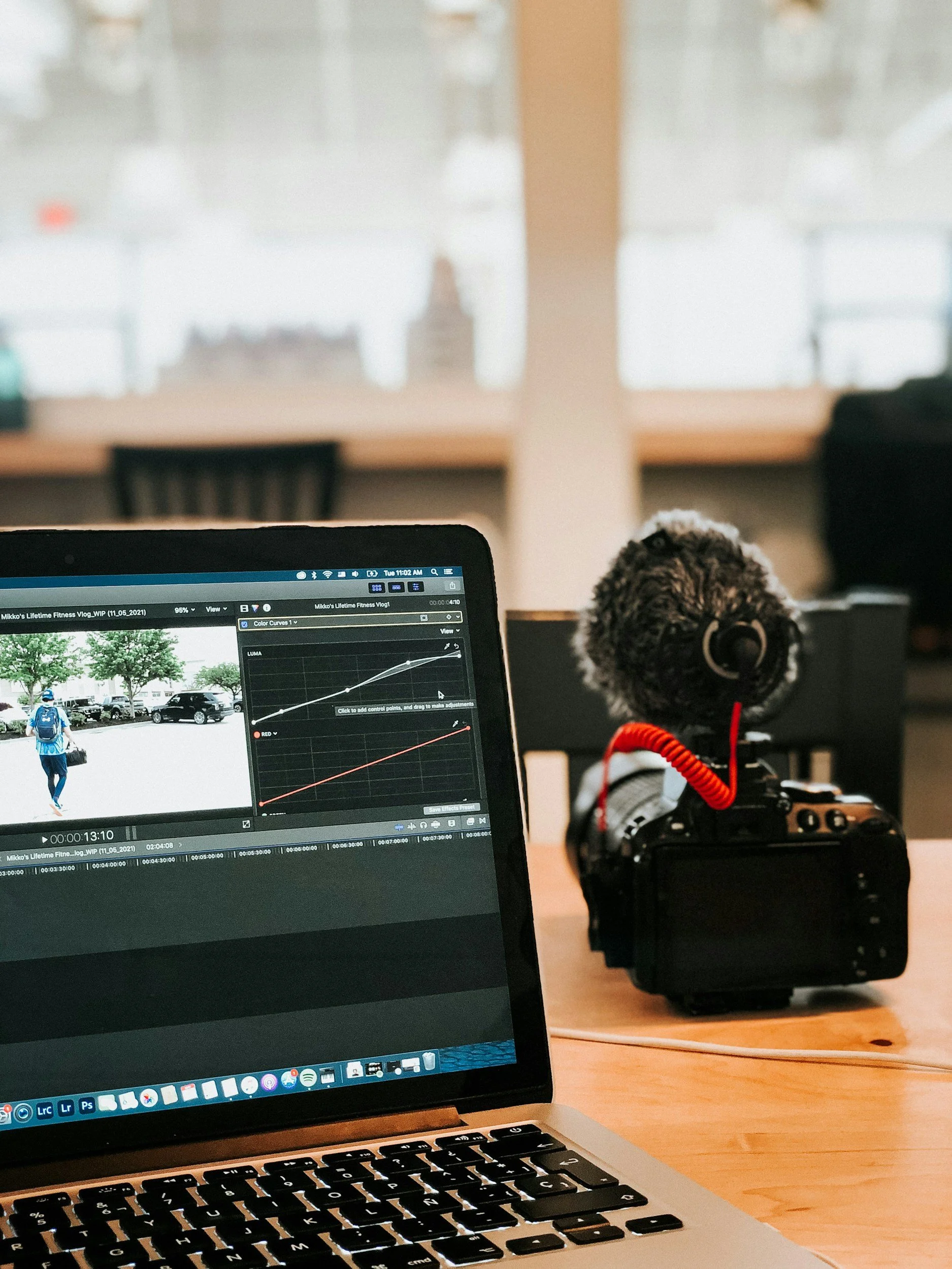 A computer screen shows a photo editing timeline with a person walking in a parking lot, wearing a blue backpack and a hat. A camera with a microphone is on the desk next to a laptop.