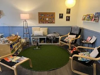 A cozy living room with a circular green rug, a blue sofa with pillows, a wooden armchair, and a small table holding magazines, with shelves of books and decorative items on the walls.