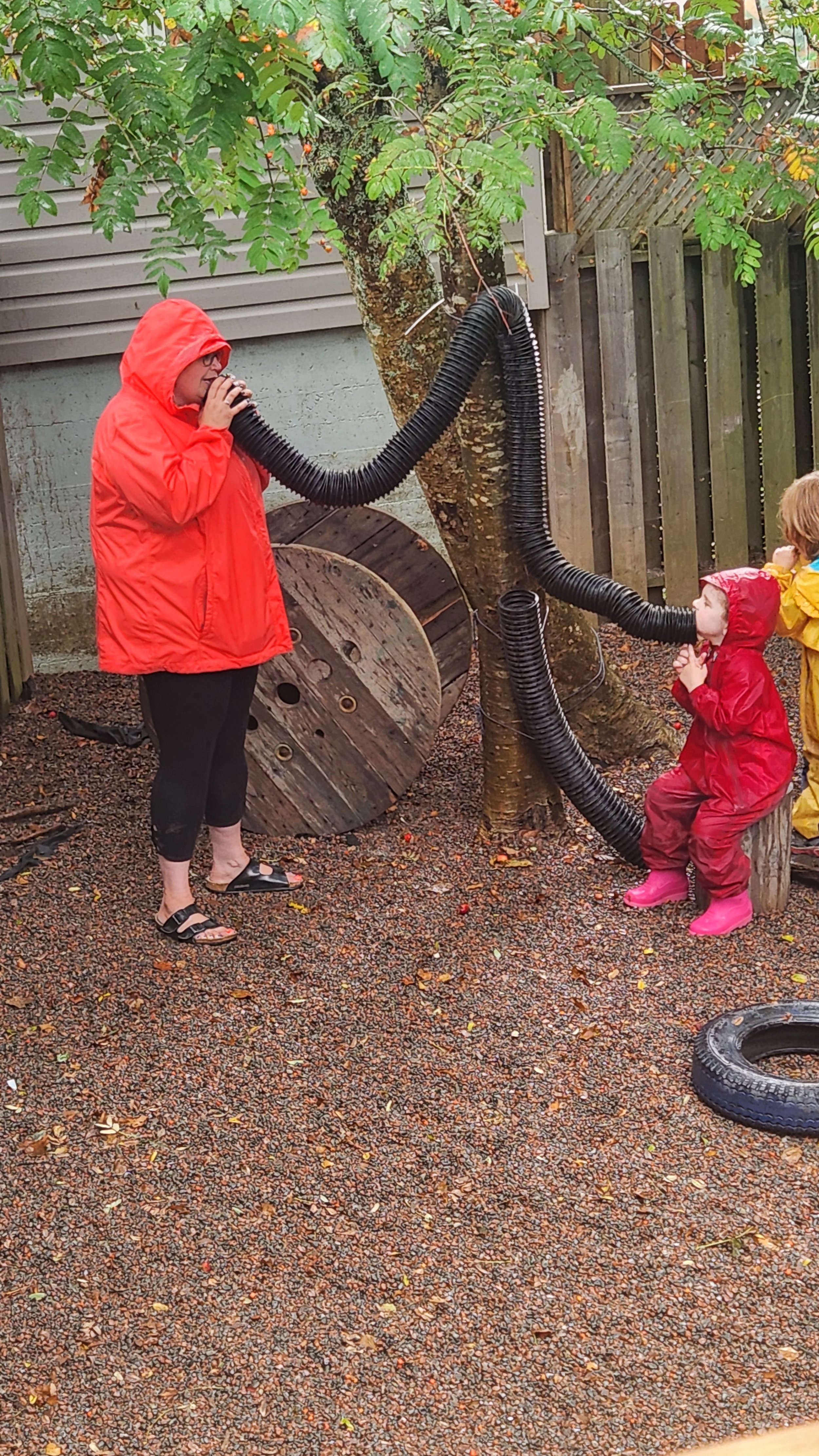 Child and woman using a large hose connected to a tree in an outdoor play area, with children in rain gear watching and a tire on the ground.