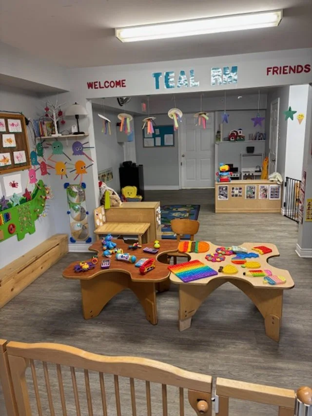 A children's play area with colorful toys on wooden tables, decorated walls with children's art, hanging paper umbrellas, and signs that read "Welcome Team Blue Friends."
