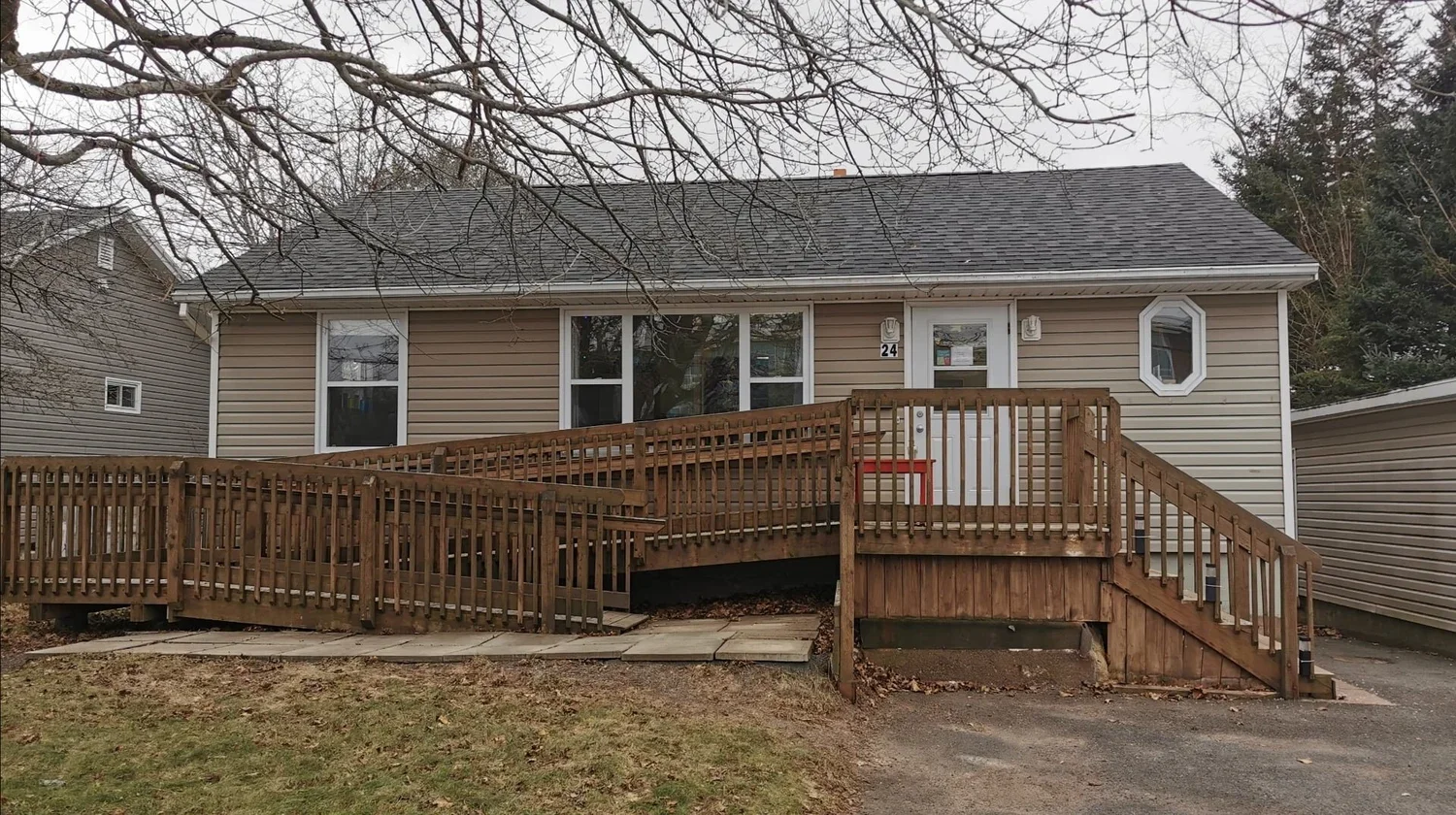 A beige house with white trim and a gray shingled roof, featuring a large window, a small octagonal window, and a white front door with house number 24. There is a multi-level wooden ramp and staircase with railings leading up to the door, and a paved driveway in front. Bare tree branches are visible above the house.