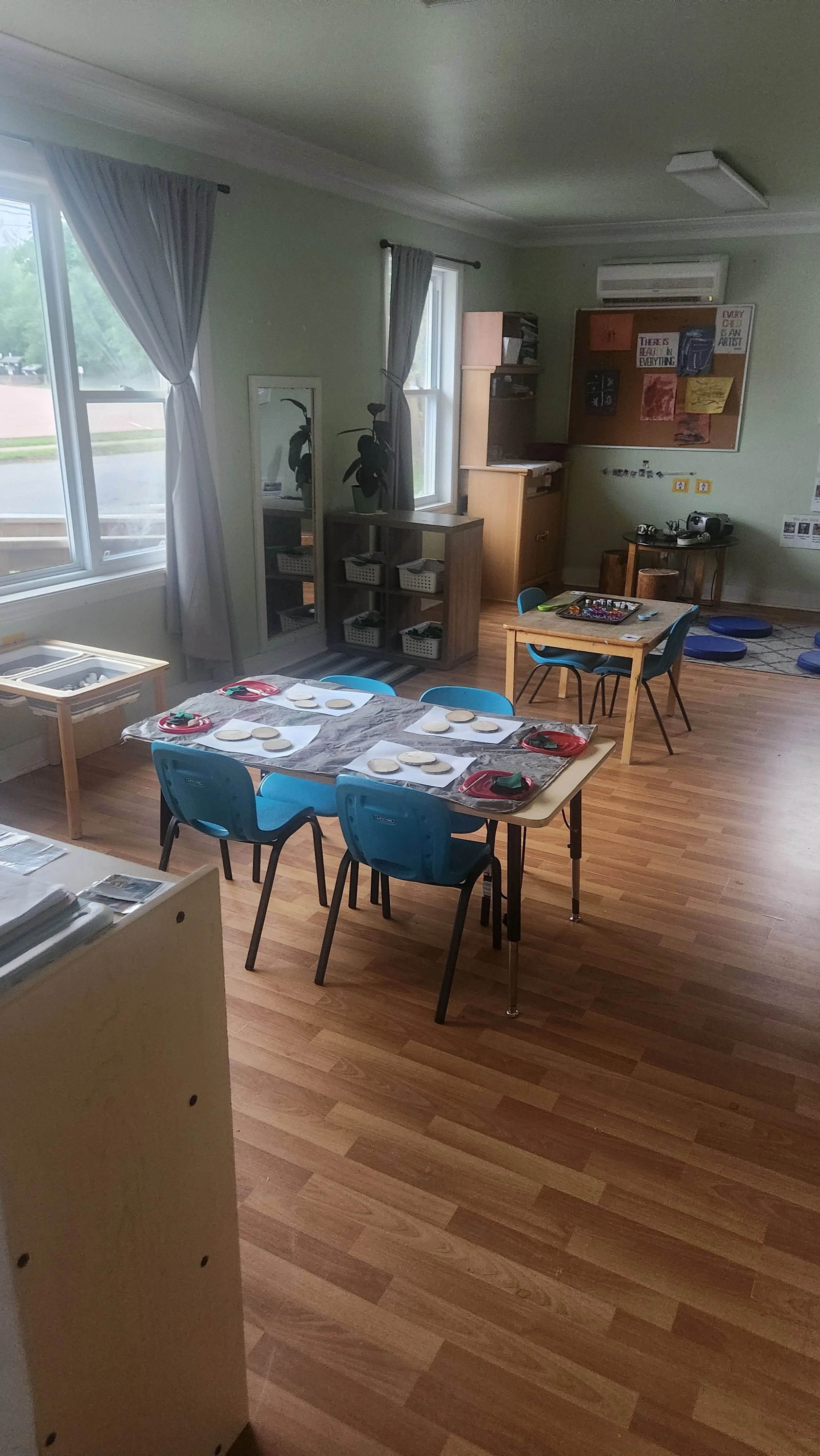 A classroom with wooden floors, blue chairs, and tables set with paper plates and utensils, suggesting a pretend play or activity setup for children.