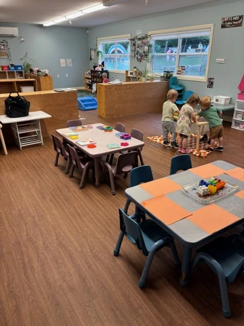 Kids playing at a table in a daycare or preschool classroom with educational toys and a play area in the background.
