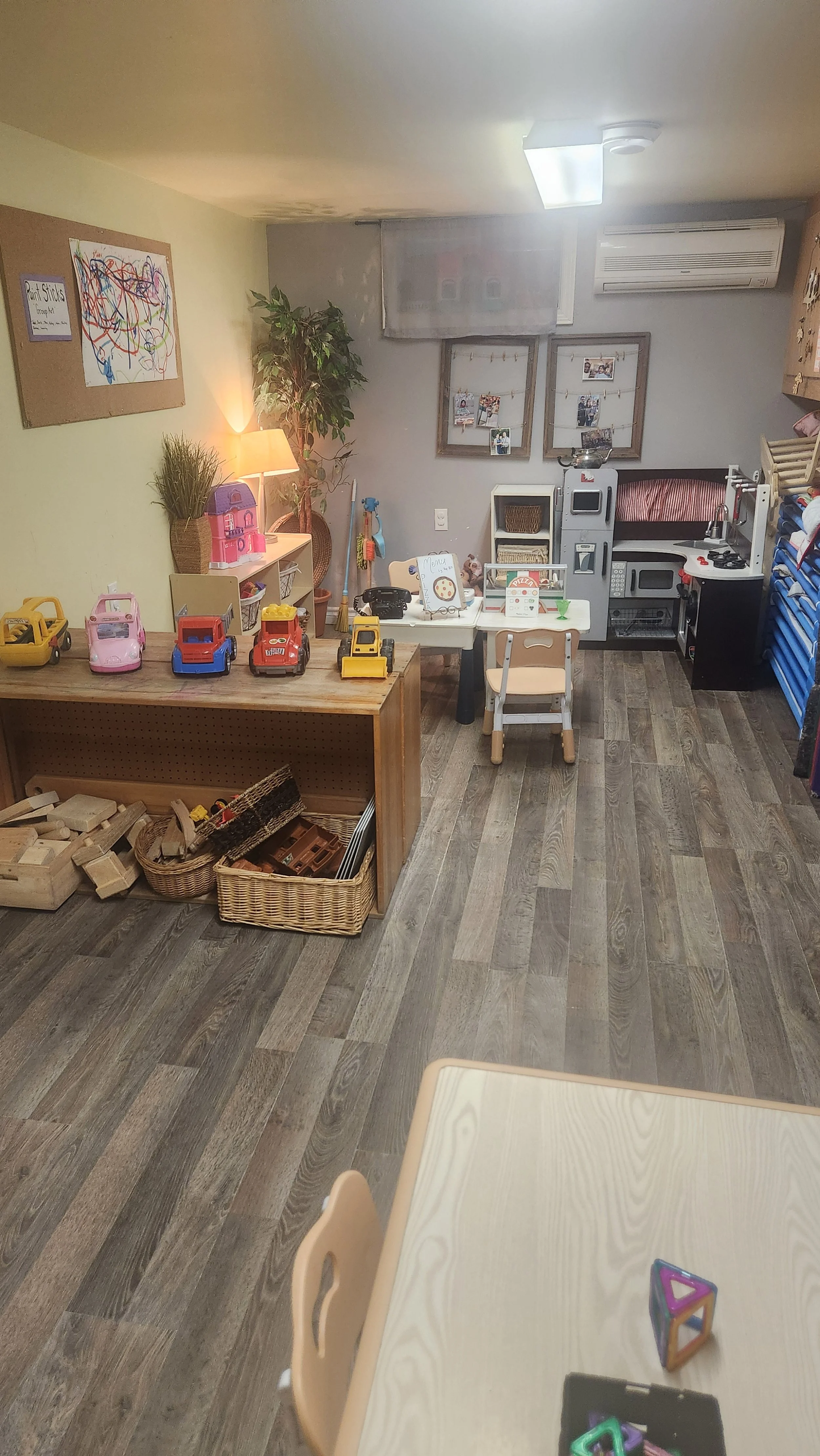 Playroom with children's toys and furniture, including a table, chairs, toy kitchen, and storage baskets, with walls decorated with framed pictures and a corkboard.