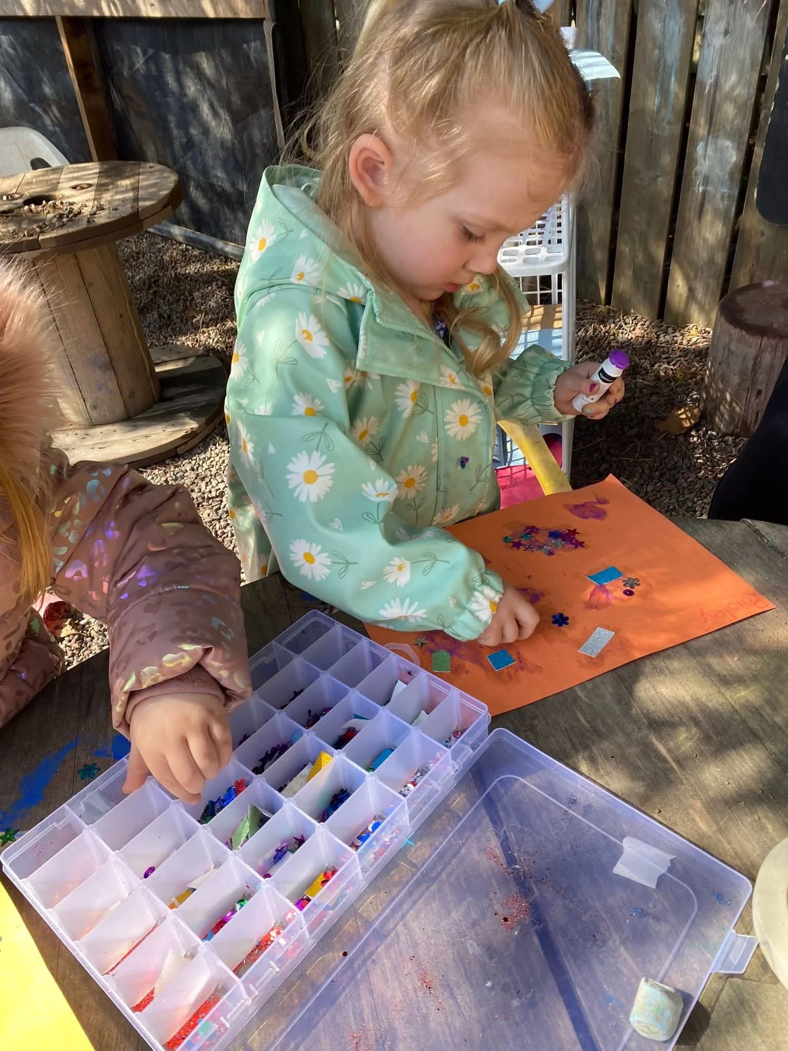 A young girl in a green hoodie decorated with daisies is working on a craft project outdoors. She is using glue and colorful sequins and stickers on an orange sheet of paper. There are containers of additional craft supplies on the table, and another child is partially visible beside her.