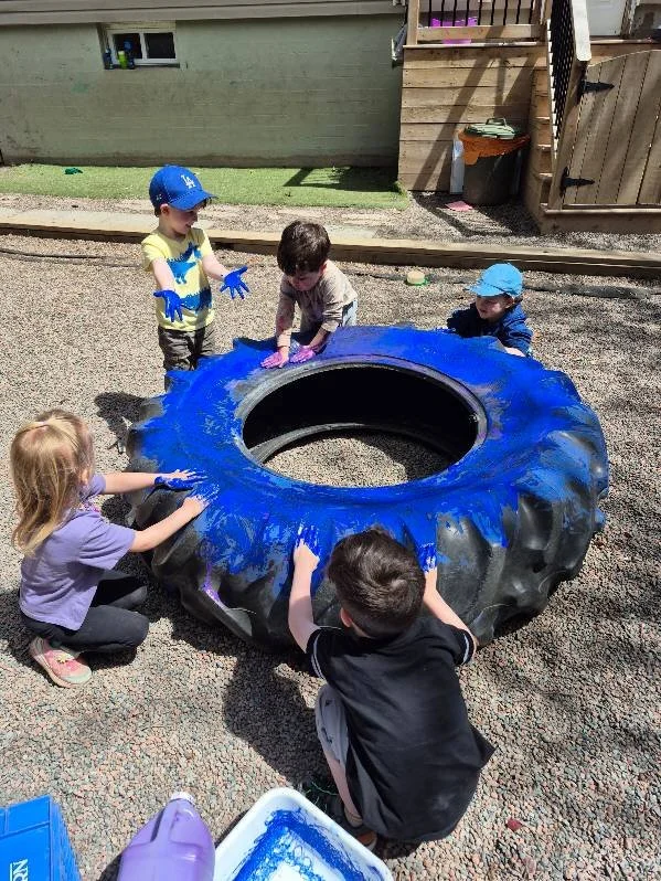Five children are painting a large tractor tire in an outdoor play area.