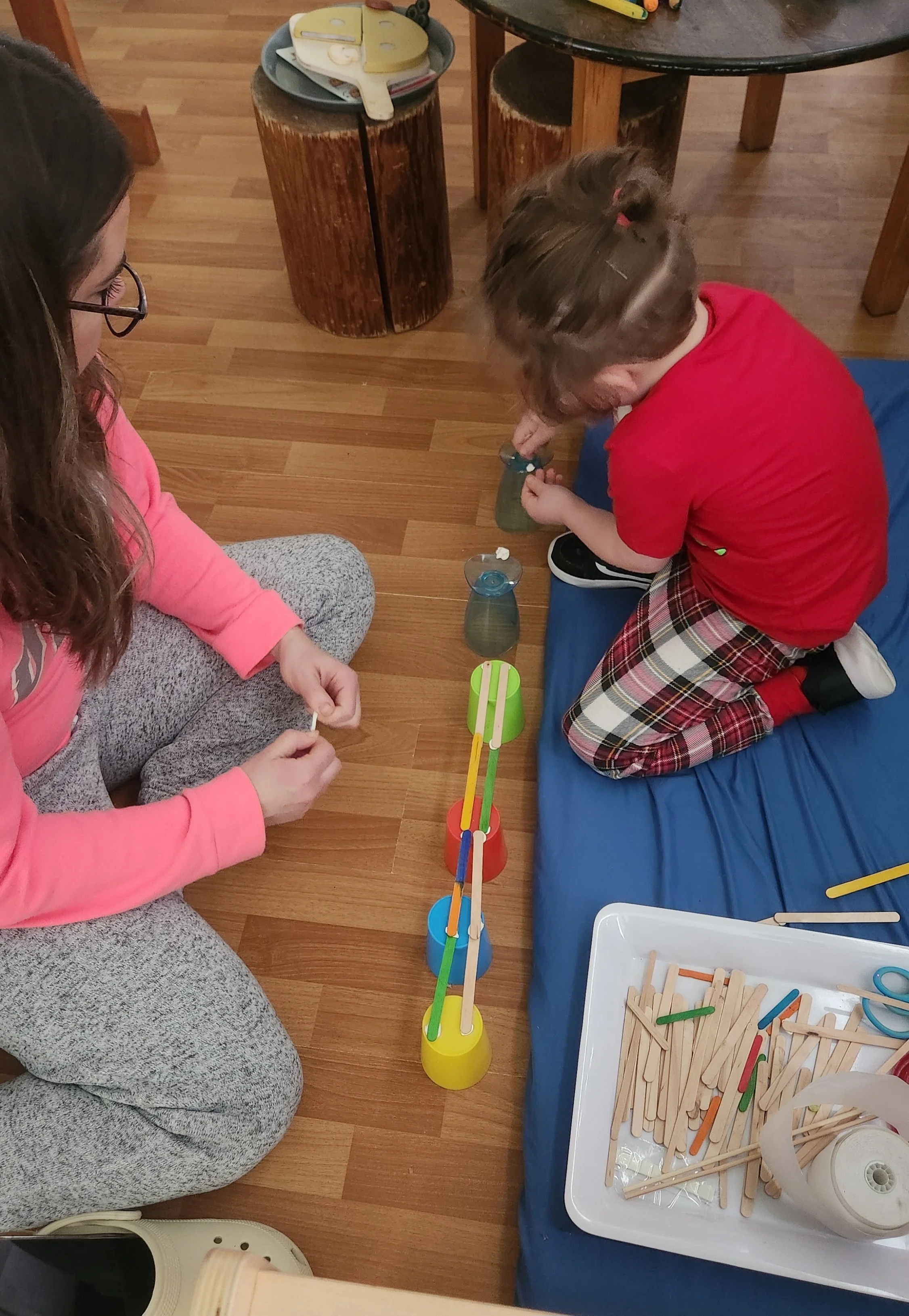 A woman and a young girl are playing a balancing game with colorful cups and sticks on a hardwood floor.