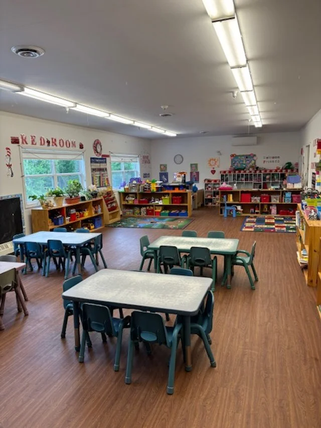 An empty preschool classroom with tables and chairs, bookshelves, and educational decorations.
