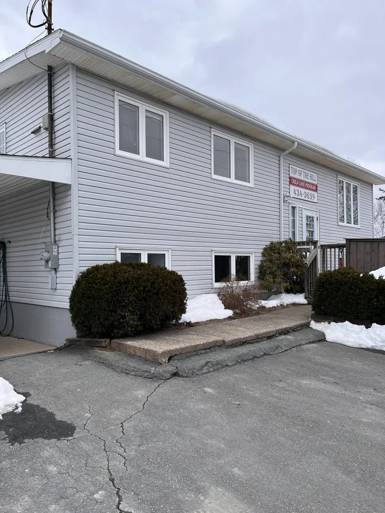 A two-story white building with siding and multiple windows outdoors in winter, with snow on the ground and bushes, and a sign for a daycare on the upper part of the building.