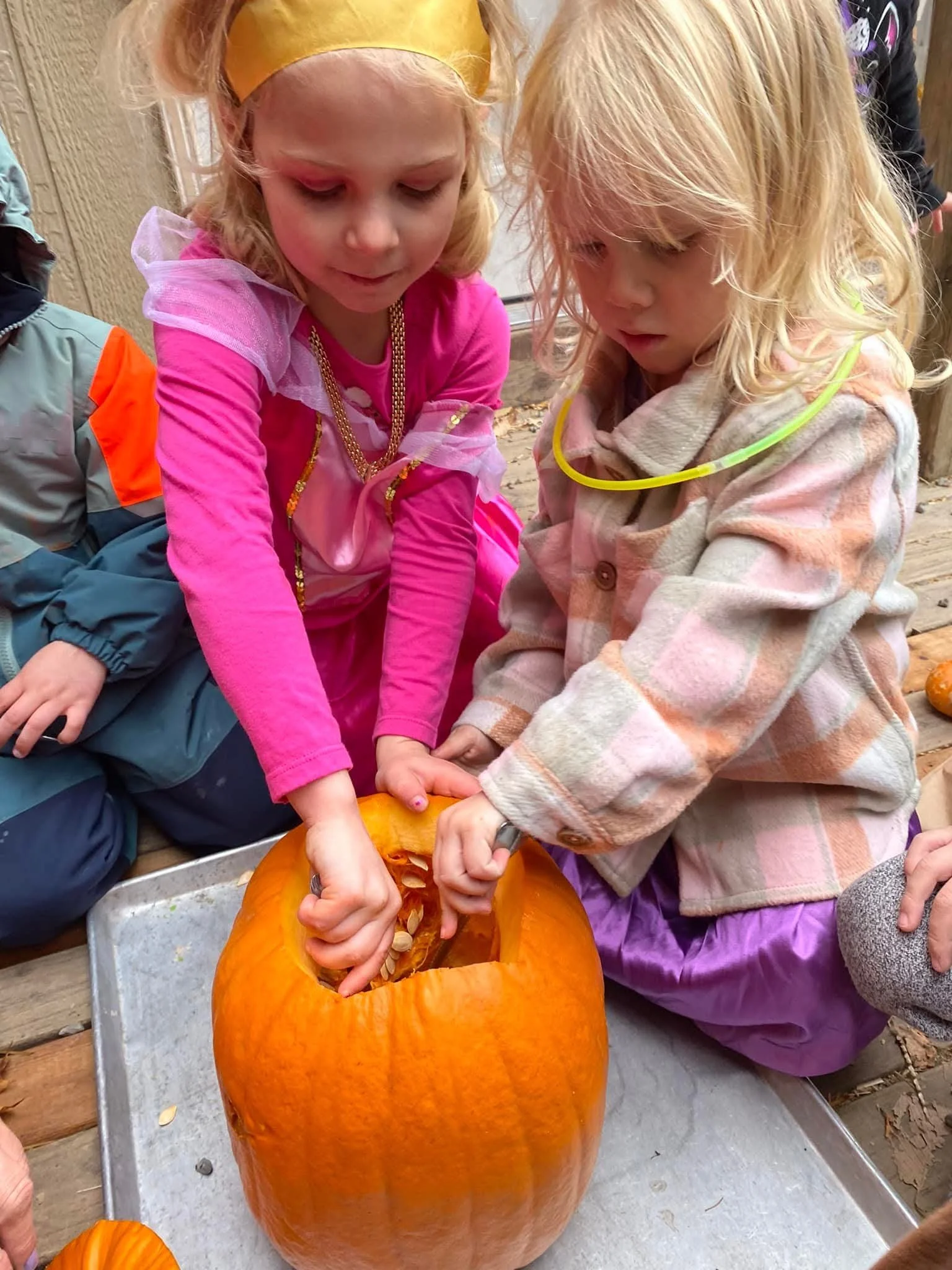 Two young girls, dressed in colorful clothing, are scooping out the insides of a large orange pumpkin during a fall pumpkin carving activity outdoors.