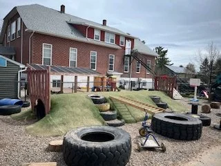 Backyard playground area with large tires, a small slide, and a grassy hill for climbing in front of a brick house.