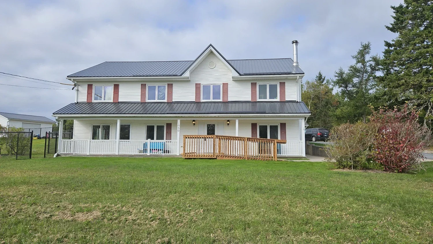 A two-story white house with pink shutters, a metal roof, and a front porch with a wooden ramp. The lawn is green with some bushes on the right and a black fence on the left. There are cars parked in the background and trees on the right side.