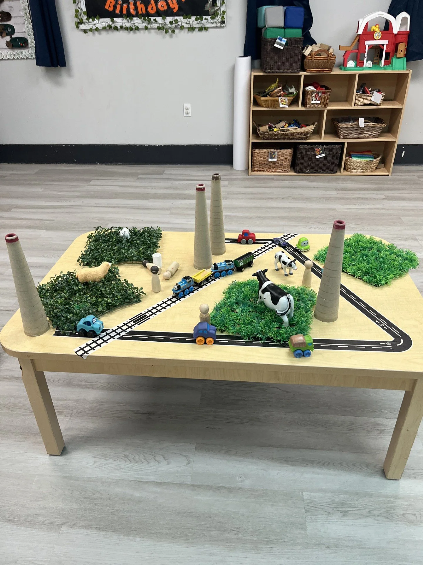 Children's play table with a miniature train set, toy animals, and green foliage, resembling a farm scene with hills and trees.