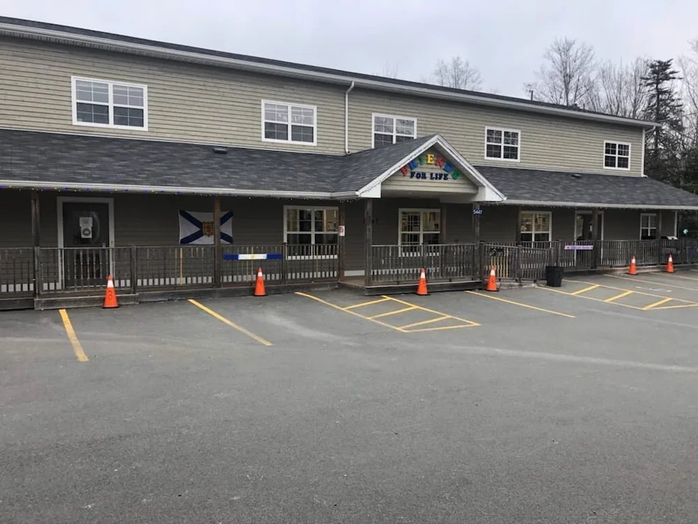 A storefront with a sign that says 'EAT FOR LIFE' decorated with colorful letters. There is a parking lot with marked spaces and orange cones in front of the building. A wooden ramp runs along the front of the building, and the windows display various flags.