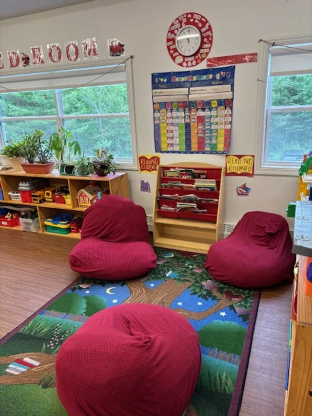 Colorful reading corner in a classroom with two red bean bag chairs, a colorful bookshelf, plants on the window sill, and a decorative rug with trees and stars.