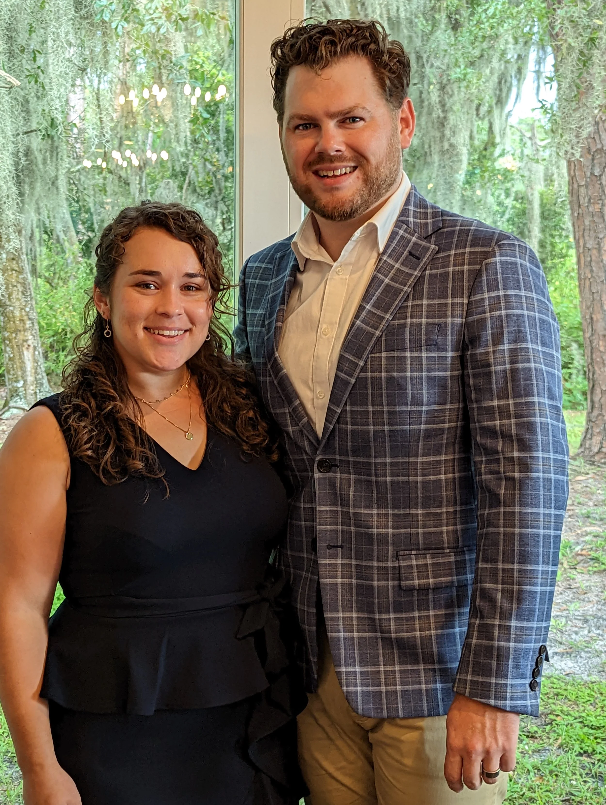 A man and woman standing together inside near a window with trees outside. The woman has curly brown hair, is wearing a black sleeveless dress, and jewelry. The man has short curly hair, a beard, and is wearing a plaid blazer, white shirt, and beige pants.