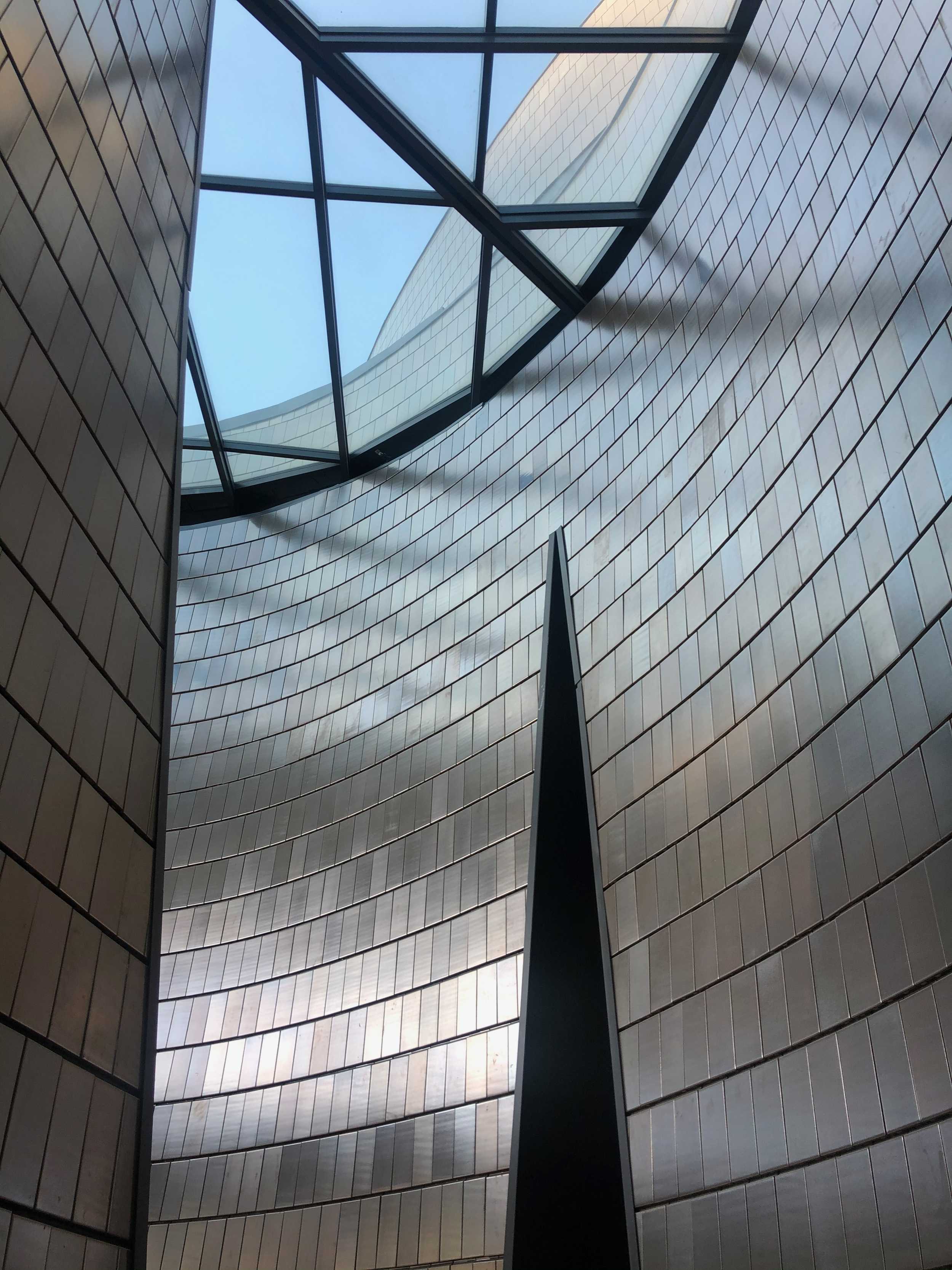 Interior view of a modern building with curved walls covered in beige tiles, view looking upward through a glass sky roof with black metal framing.