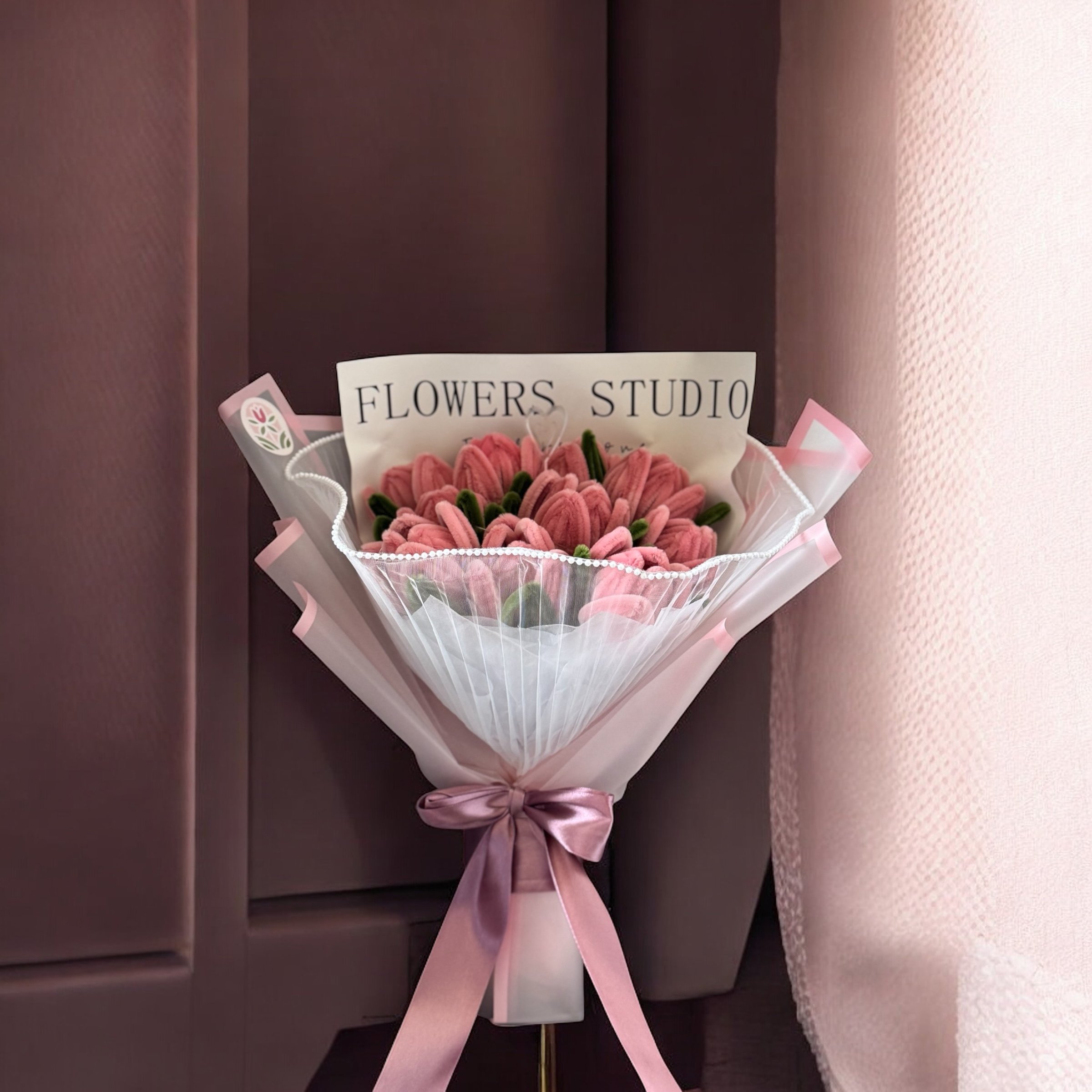 Pink flower bouquet with a sign reading 'Flowers Studio' wrapped with pink ribbons and placed against a dark and light pink background.