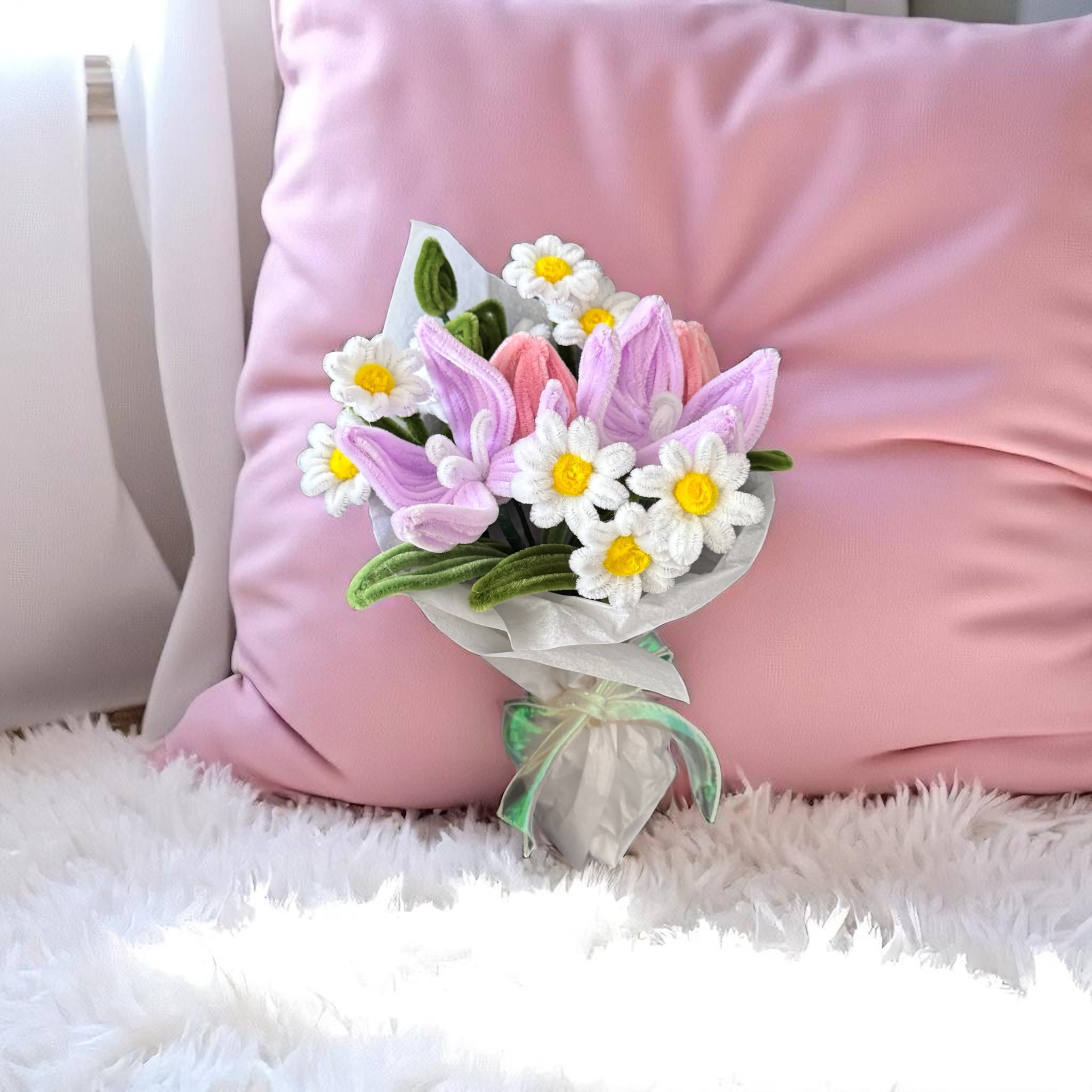 A bouquet of artificial pink tulips, white daisies with yellow centers, and green leaves placed against a pink pillow on a fluffy white surface.