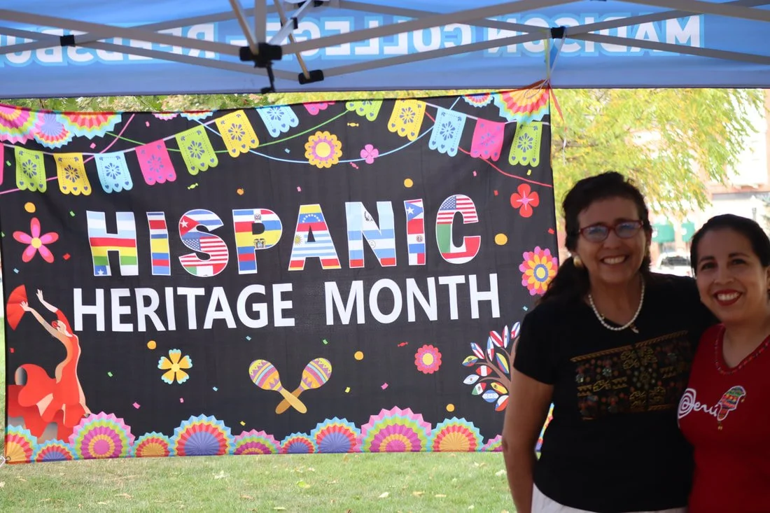 Two women standing in front of a colorful sign that reads 'Hispanic Heritage Month,' decorated with festive patterns, flowers, and flags, at an outdoor event.