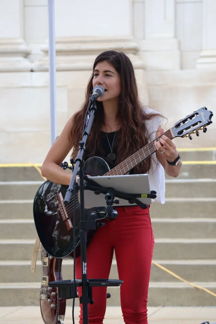Young woman with long dark hair playing guitar and singing into a microphone on steps outside, wearing red pants and a white top.