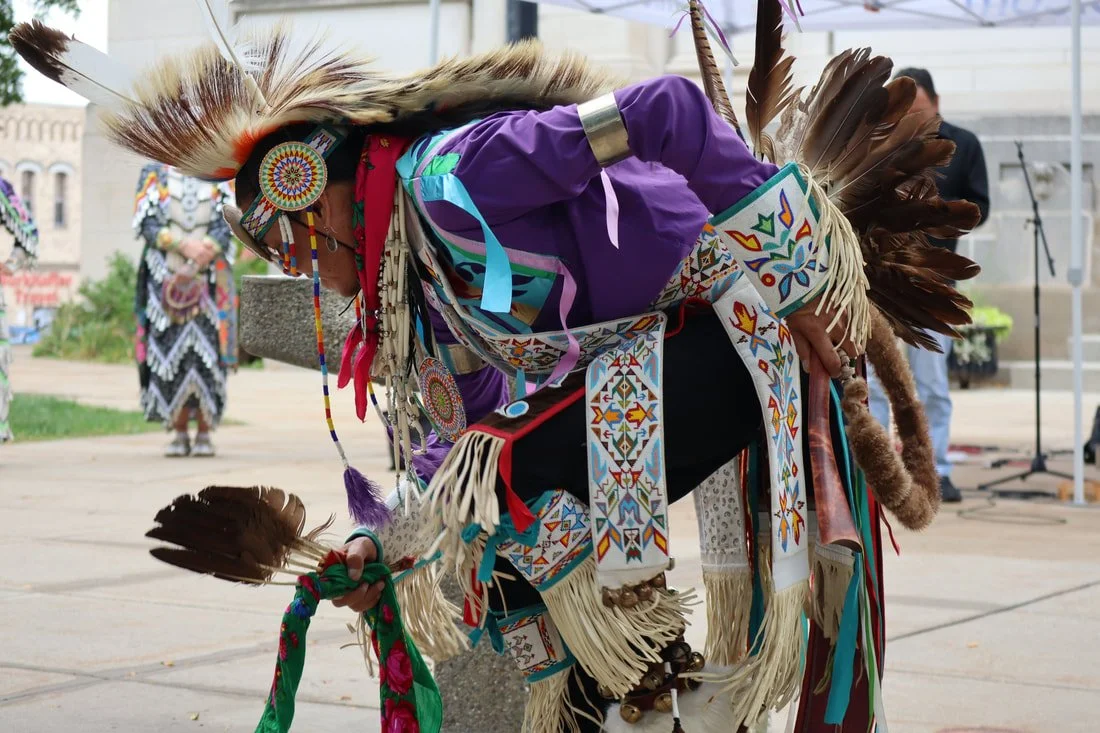 Person dressed in traditional Native American regalia bowing at a cultural event, wearing a feathered headdress, colorful beadwork, and decorated clothing, outdoors in an urban area.