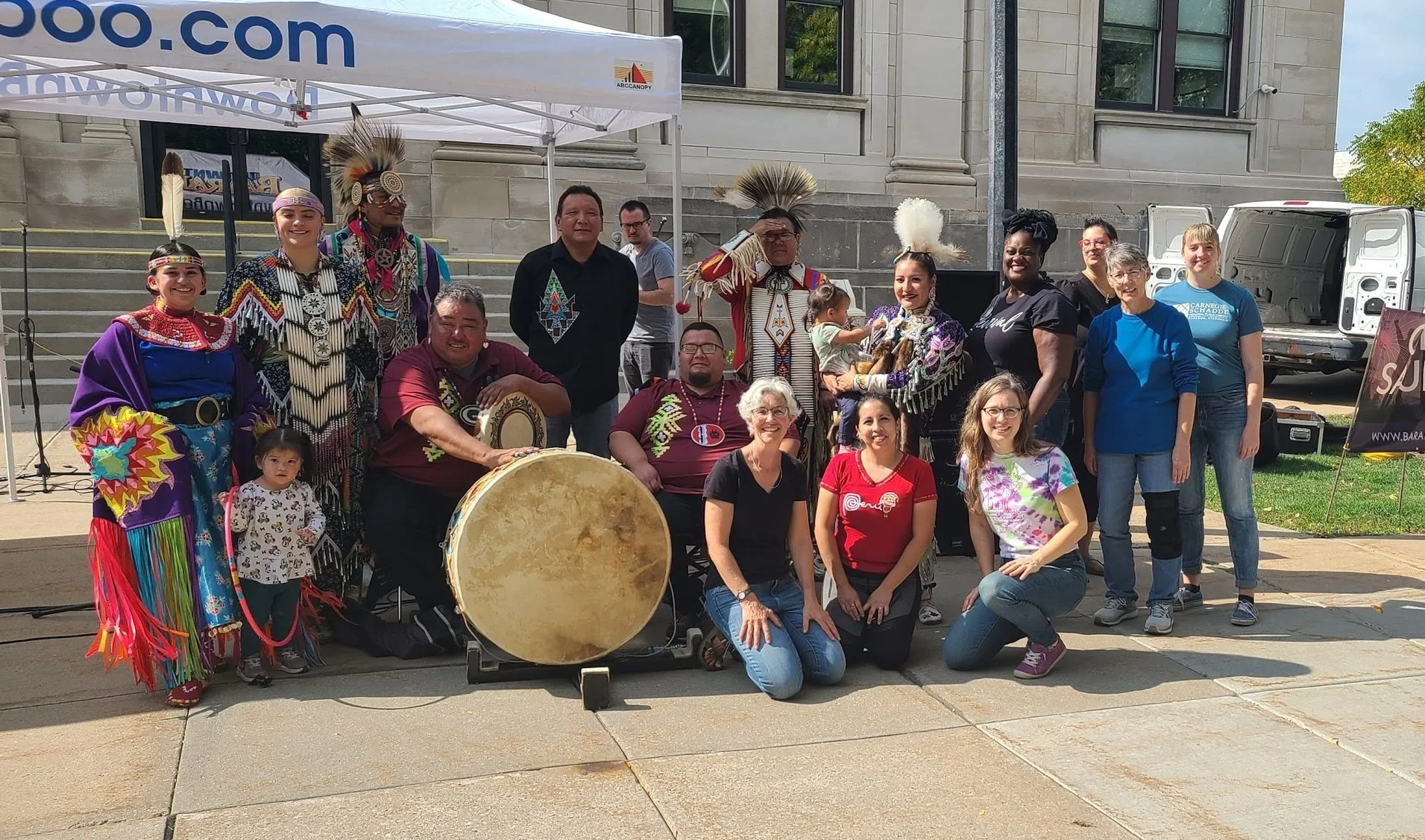 Group of people, including Indigenous performers in traditional attire, some with feathered headdresses, holding drums and musical instruments, gathered outdoors for a cultural event in front of a large building, with a white tent and other participants standing behind.