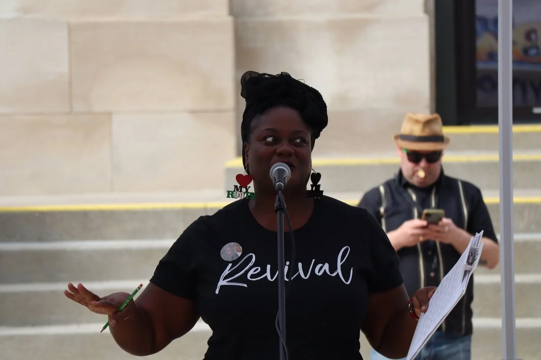 African American woman speaking into a microphone outdoors, wearing a black t-shirt with 'Revival' written on it and colorful earrings, with steps in the background. A man wearing a hat and sunglasses, using his phone, stands behind her.