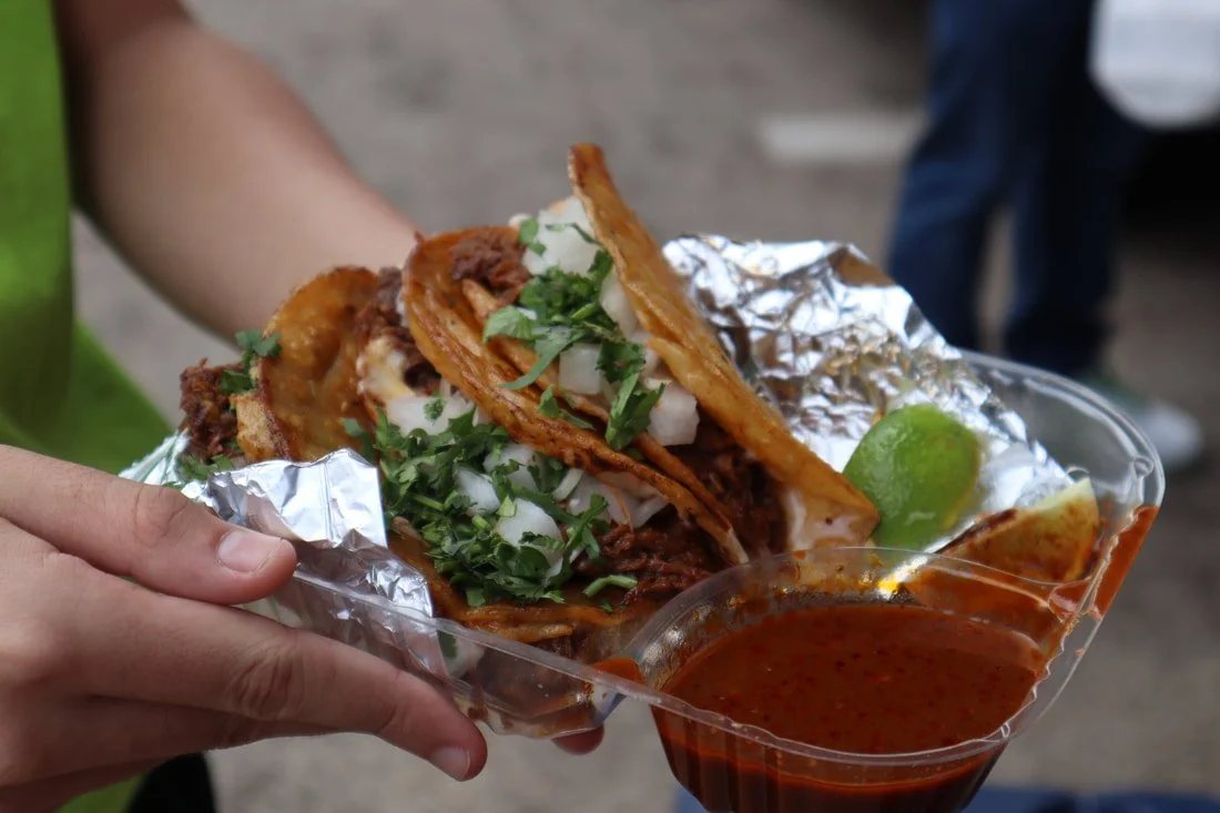 Person holding a plastic tray with two tacos filled with shredded meat, chopped onions, cilantro, and a wedge of lime, alongside a cup of red salsa.