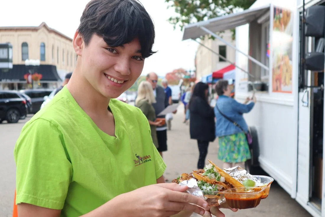 Smiling young man in bright green shirt holding a food tray at an outdoor food festival, with food and people in the background.