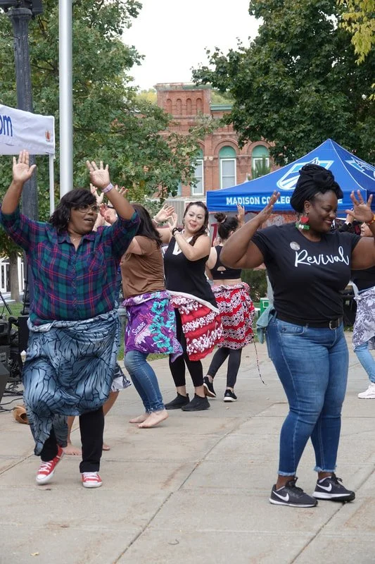 Women dancing outdoors at a cultural event, some wearing traditional skirts and tops, under a blue tent in front of a brick building.