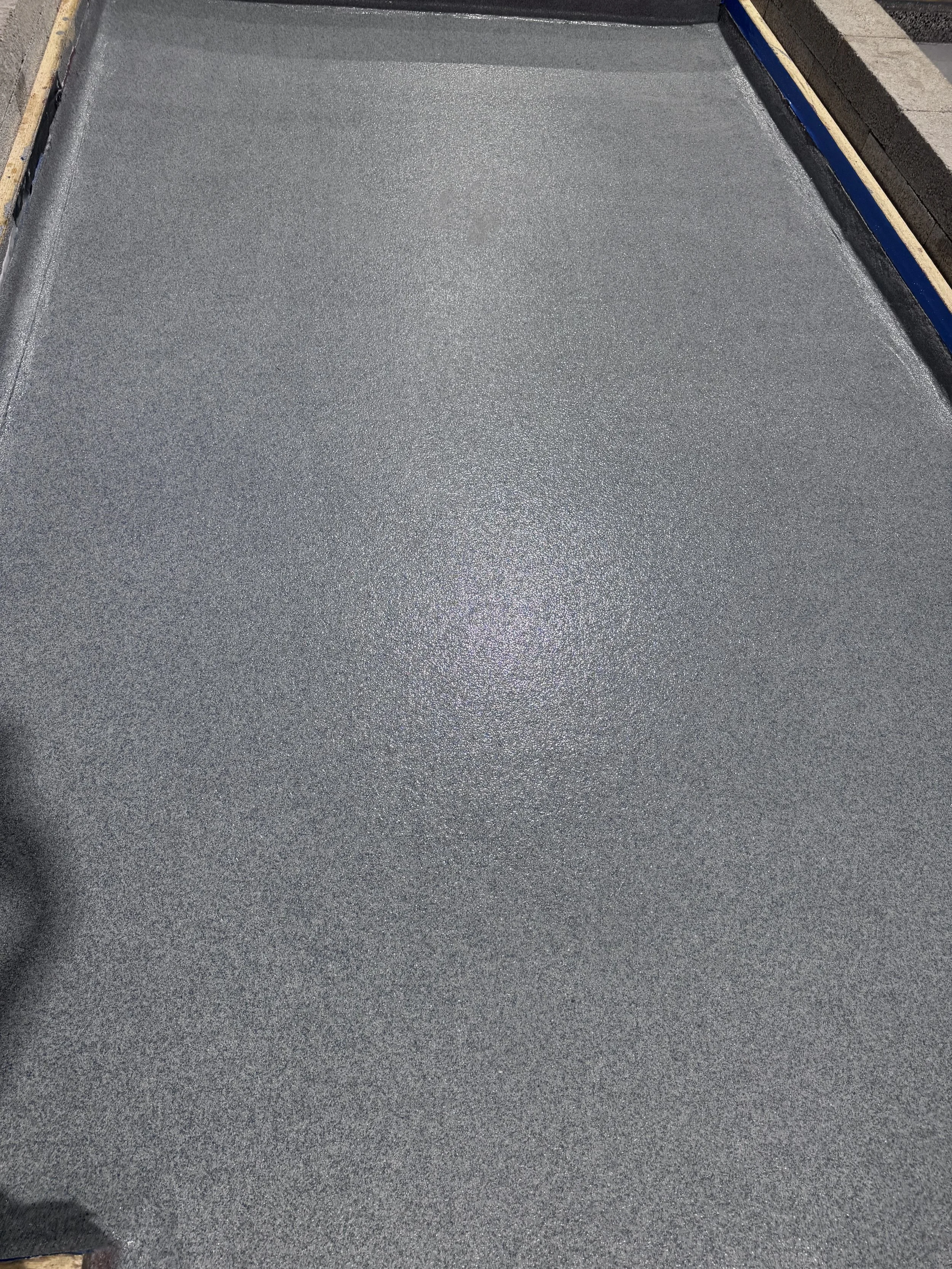 A freshly laid concrete slab floor with a textured surface, surrounded by construction materials and partially installed flooring at the edges.