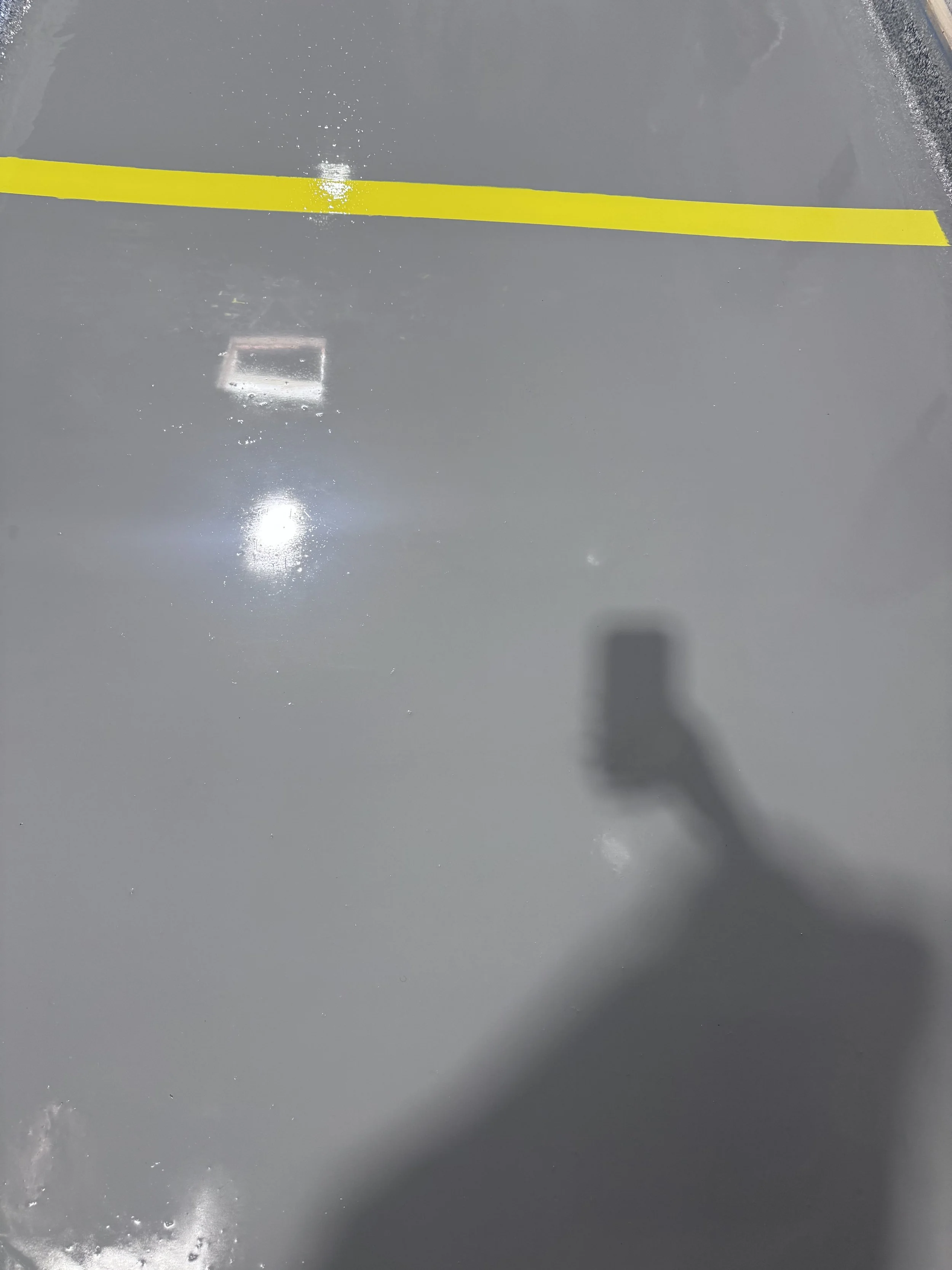 The image shows a smooth, shiny gray floor with a yellow line painted on it, and the reflection of overhead lights and a person holding a camera or phone.