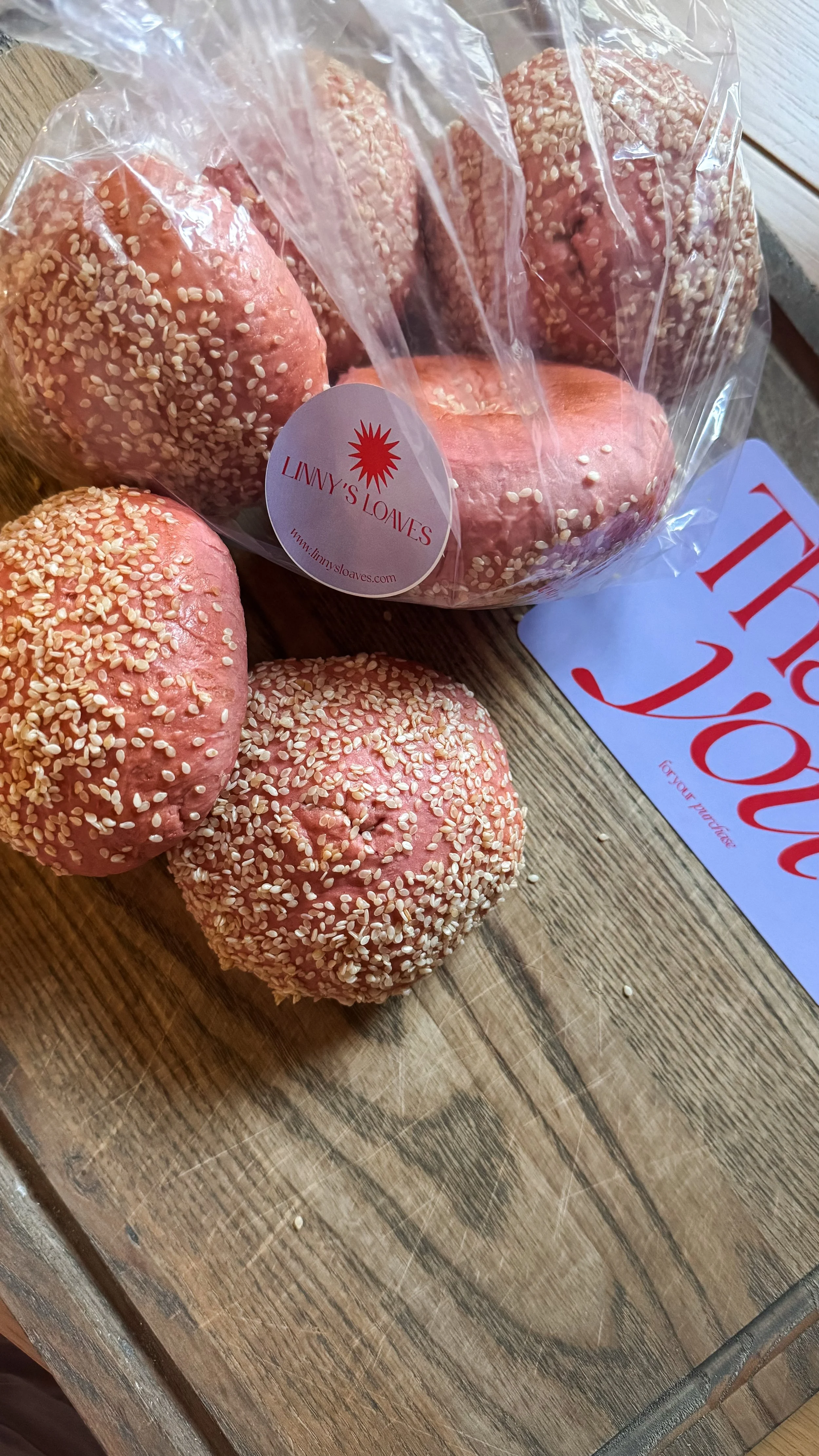 Pink bagels topped with white sesame seeds on a wooden surface, with a clear plastic bag and a 'Thank You' card nearby.