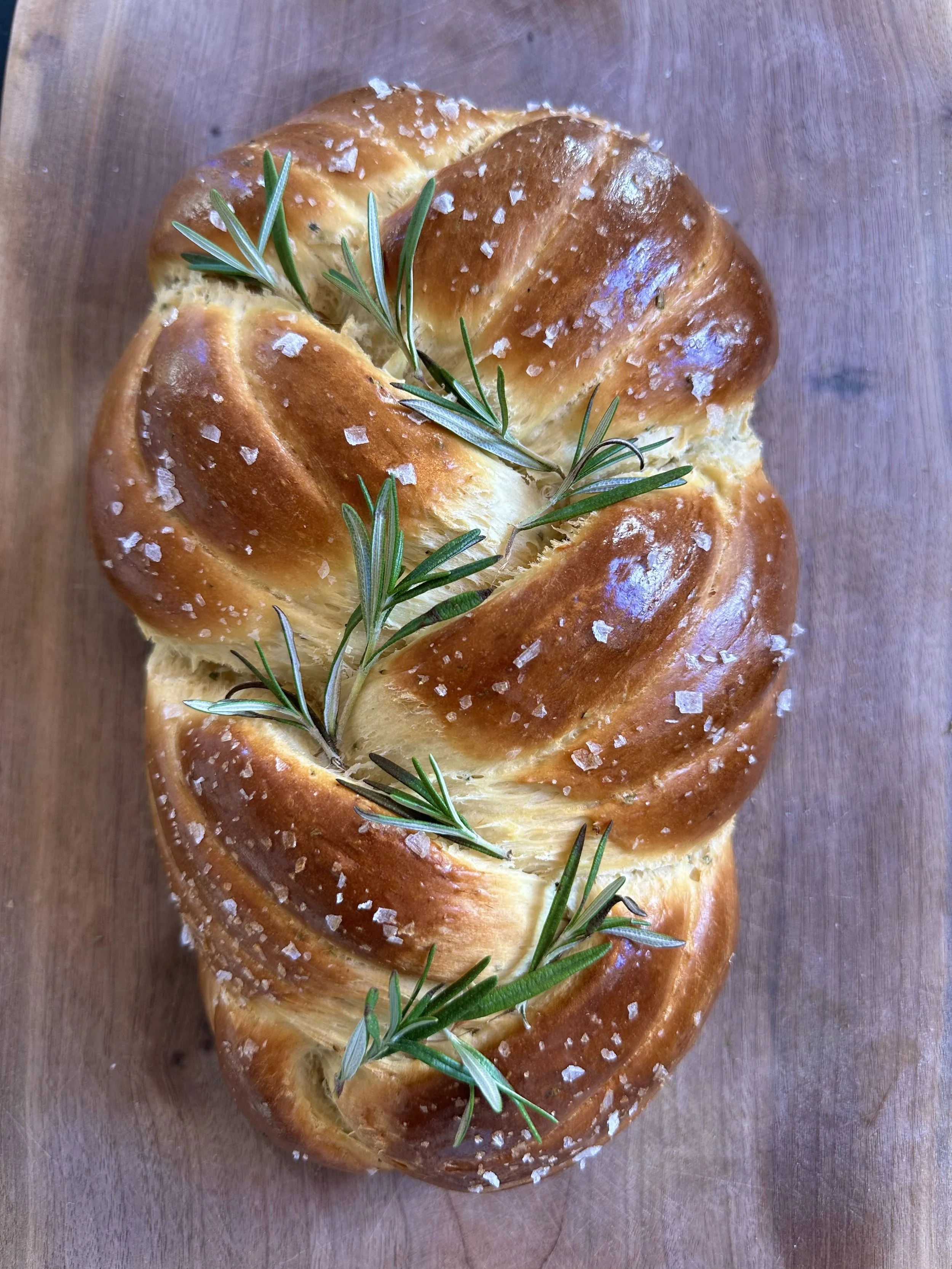 Freshly baked twisted bread loaf topped with coarse salt and garnished with sprigs of rosemary, sitting on a wooden surface.