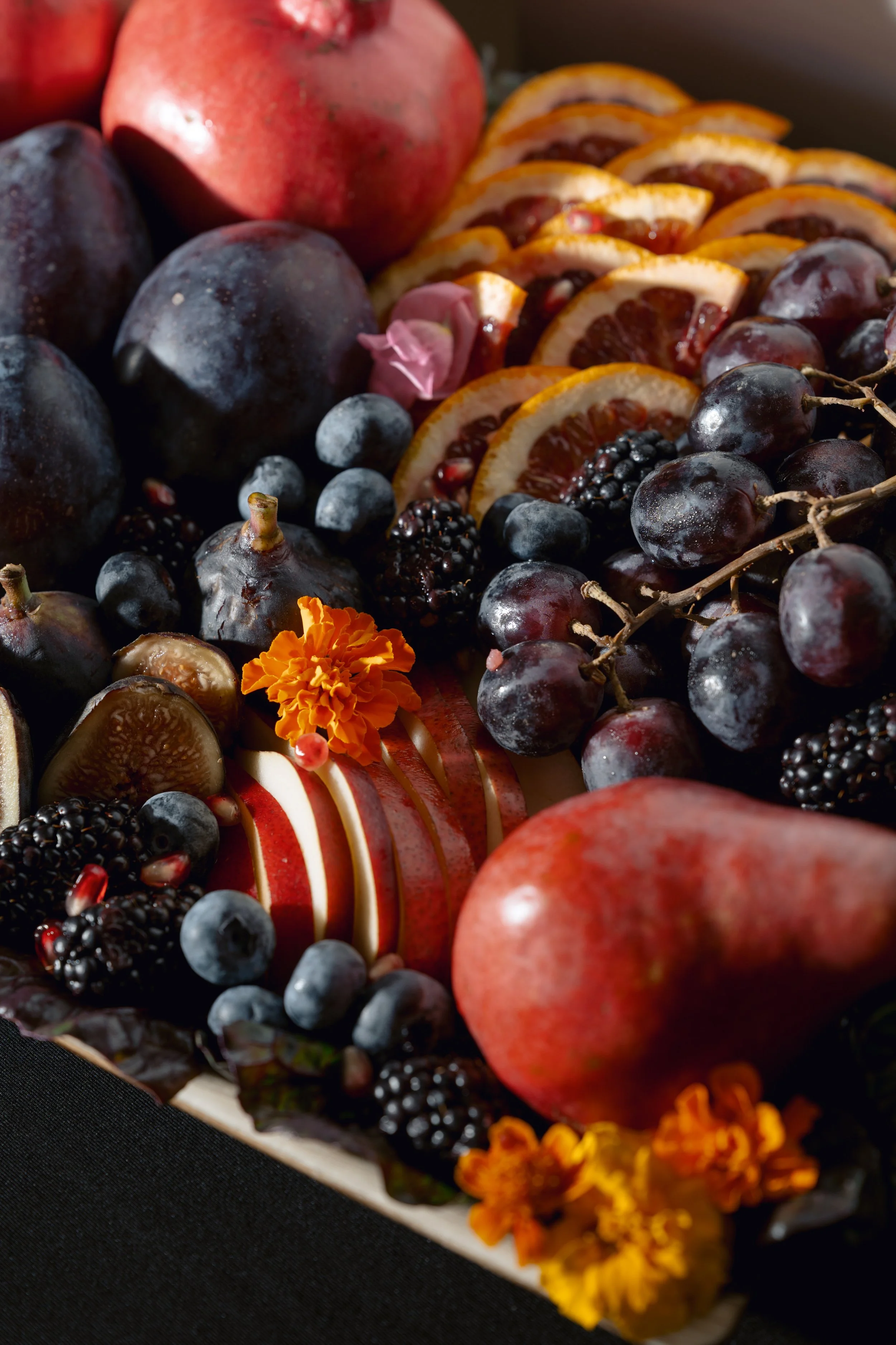 Fresh assorted fruits including pomegranates, black grapes, blueberries, figs, blackberries, and sliced blood oranges on a platter, decorated with marigold flowers.