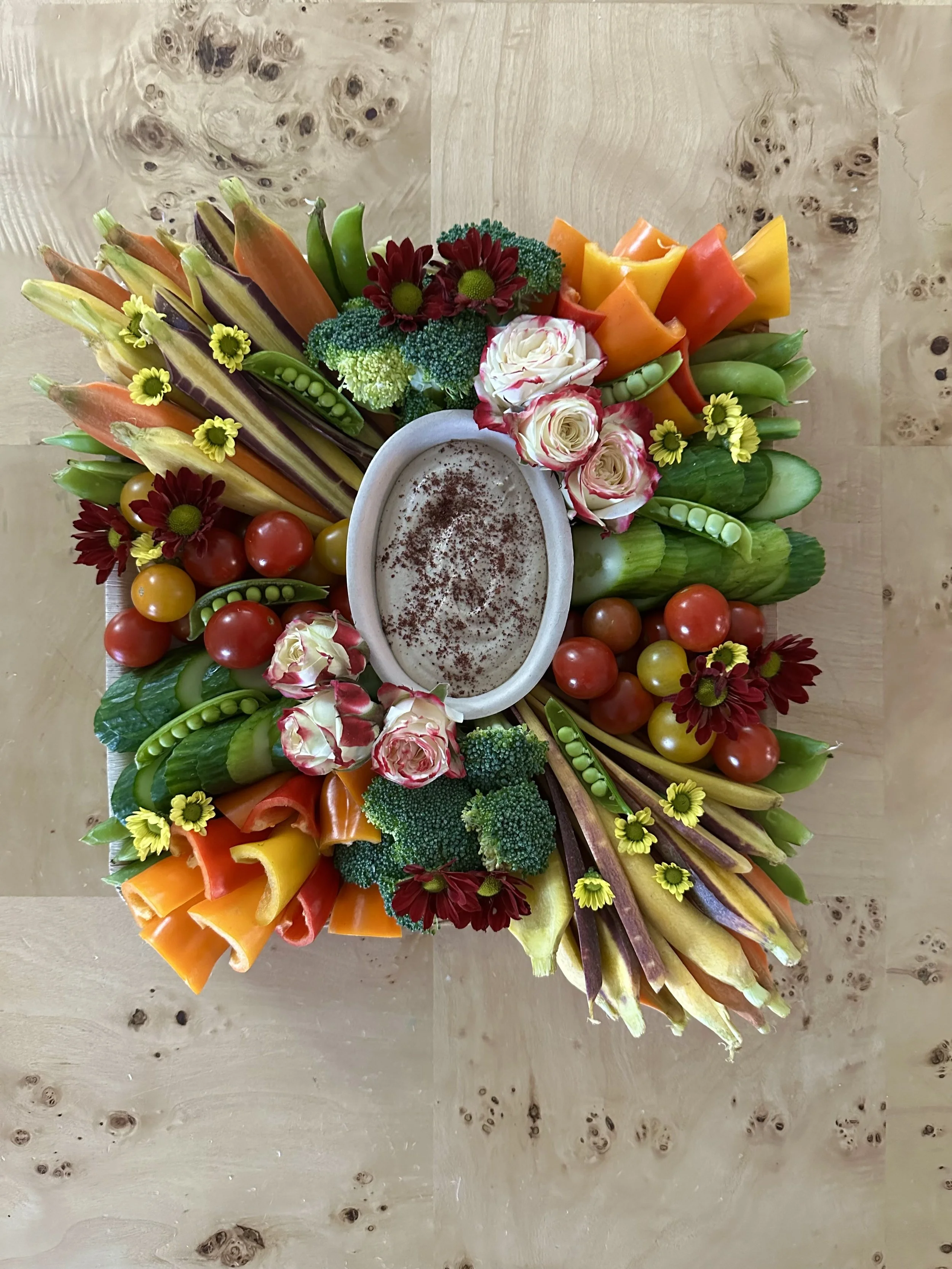 Colorful vegetable platter arranged in a circle around a bowl of dip with a sprinkle of spices, including cherry tomatoes, broccoli, carrots, cucumbers, radicchio, and assorted flowers, on a wooden surface.