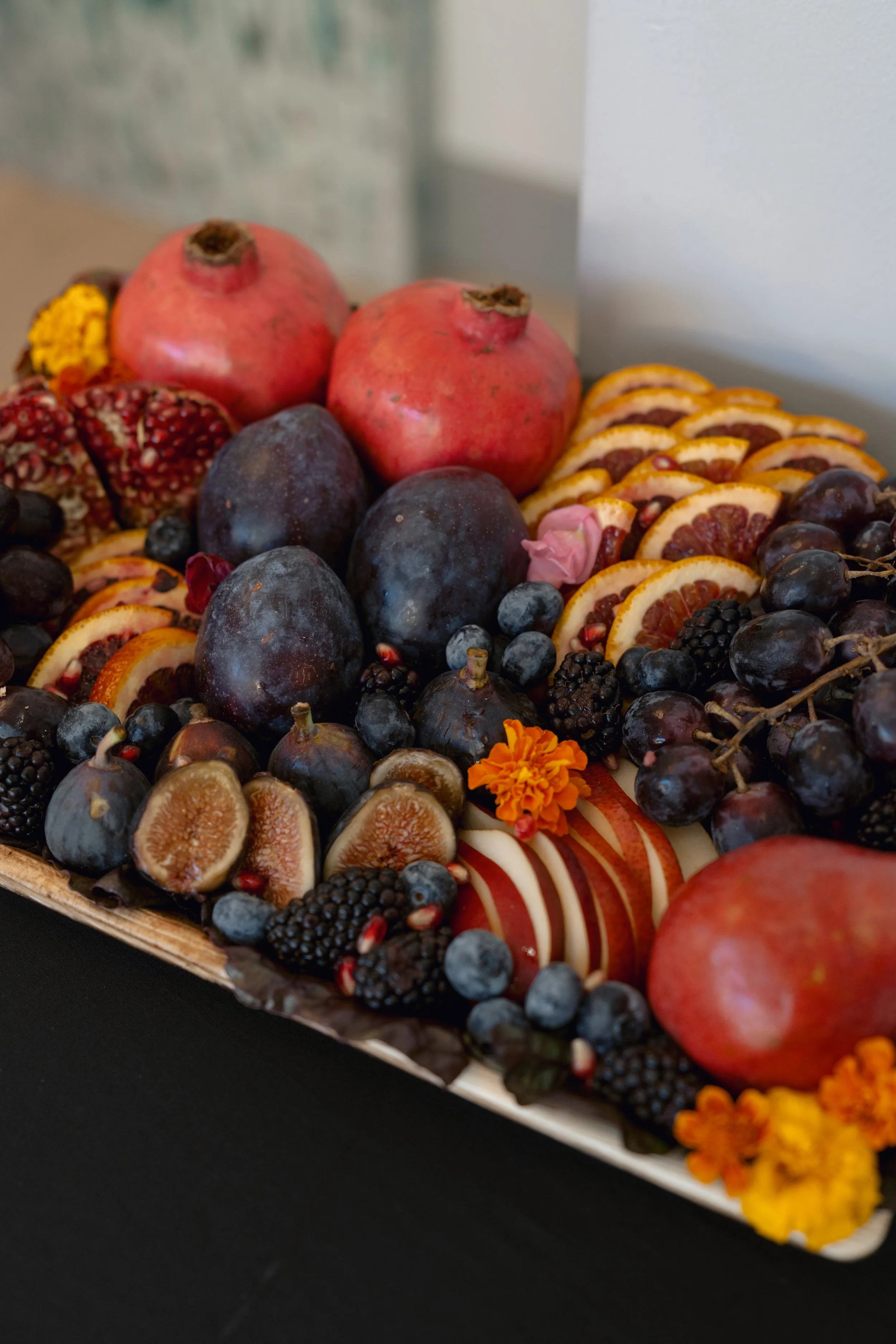 Tray of assorted fresh fruits including pomegranates, figs, blackberries, blueberries, deep purple grapes, and slices of apple and blood orange garnished with small flowers.