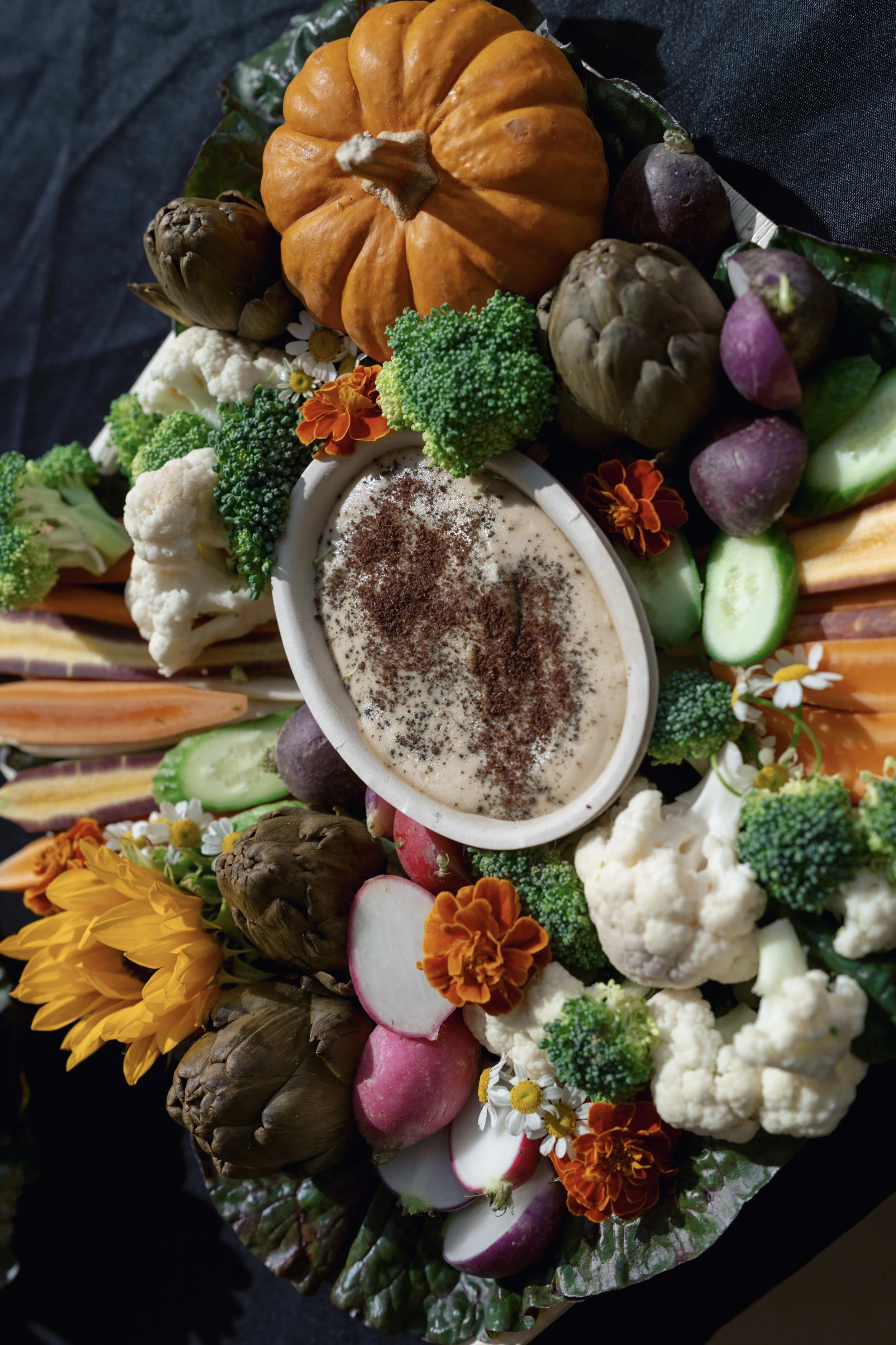 A colorful vegetable platter with broccoli, cauliflower, radishes, carrots, artichokes, cucumber, and a small pumpkin, with a bowl of dip in the center.