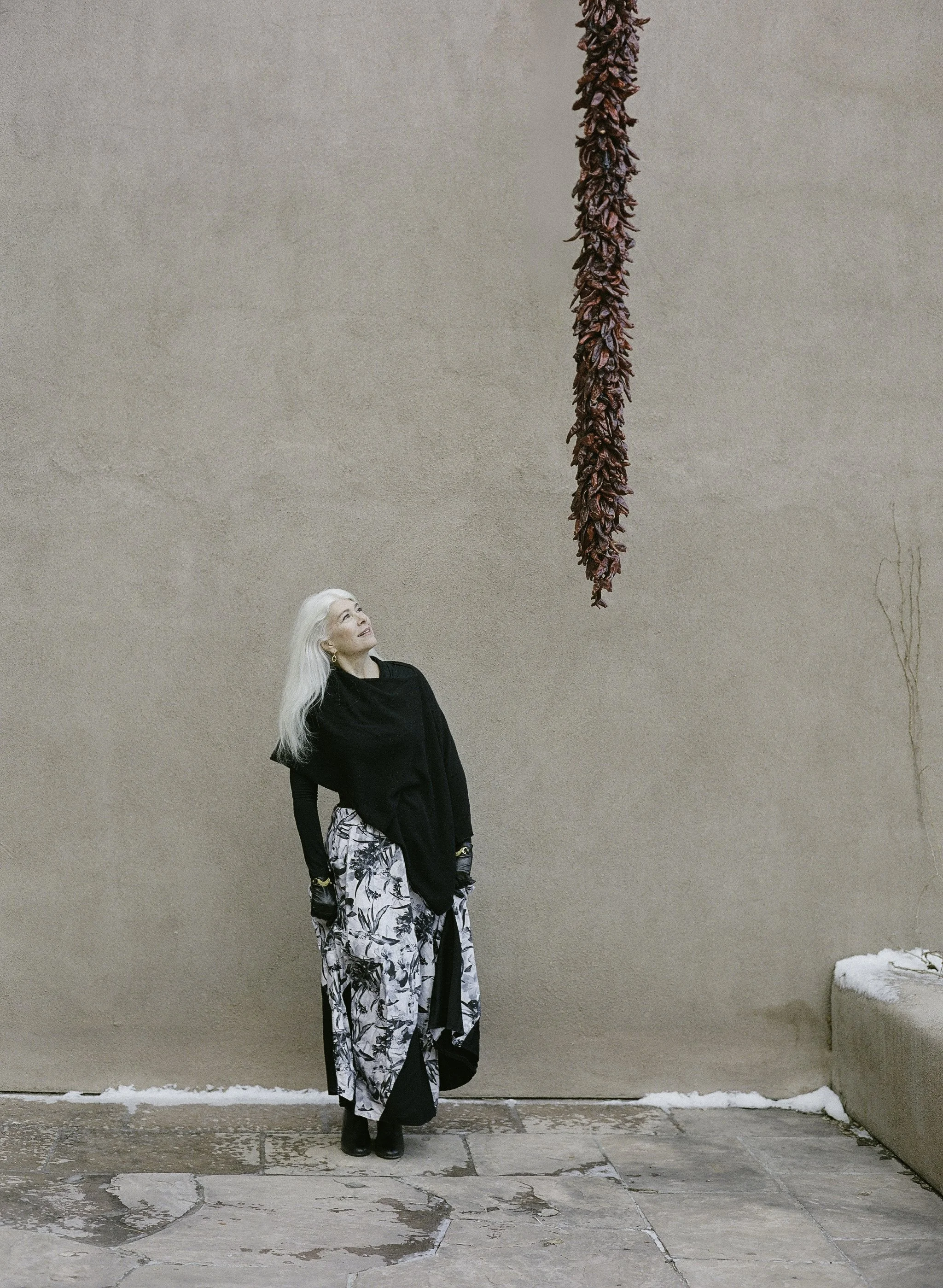 Editorial portrait on film — silver-haired woman in black wrap and floral skirt looking up at dried chilli ristra against adobe wall, New Mexico, by Radostina Boseva
