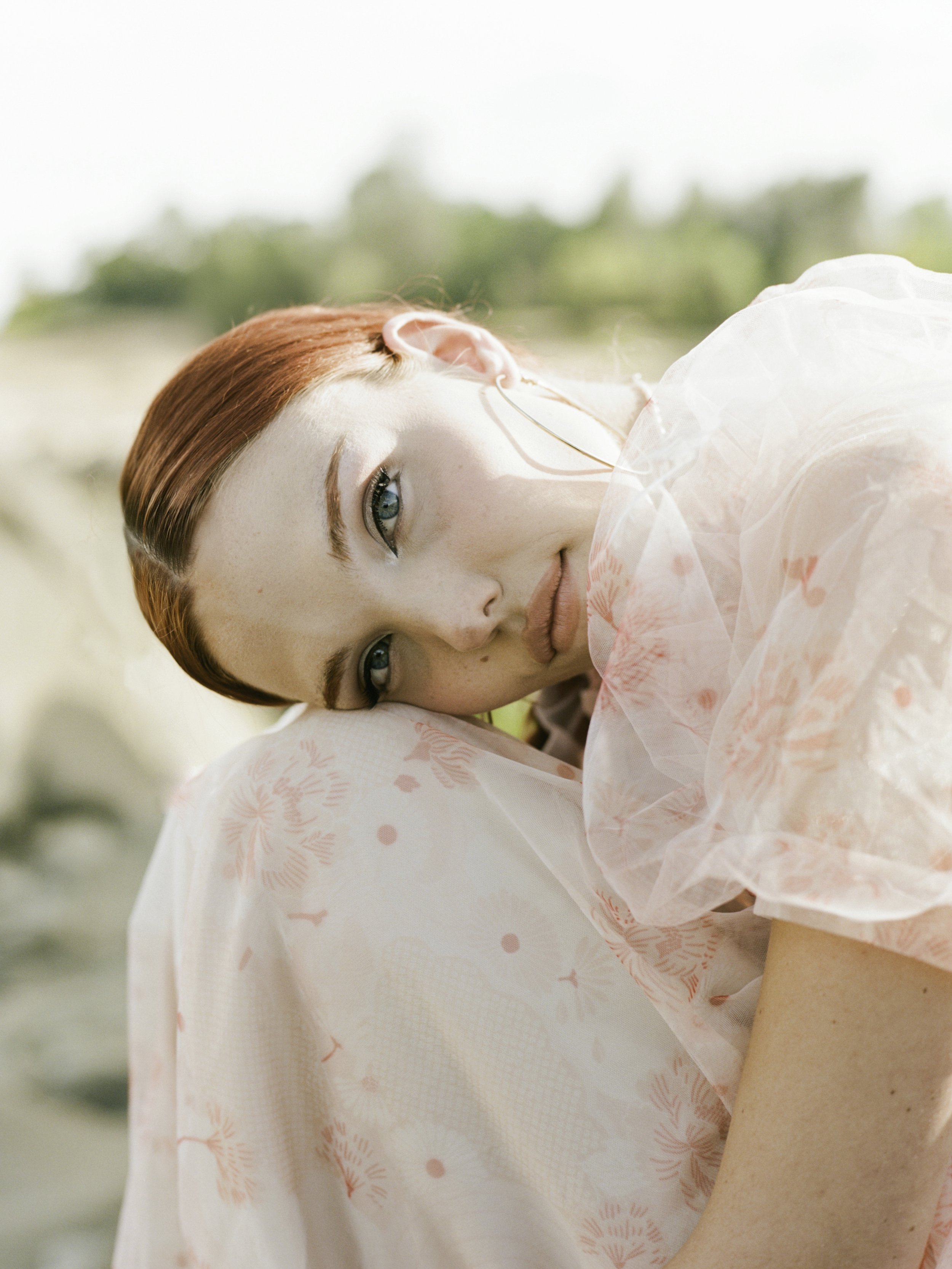 Close editorial film portrait of red-haired woman in pink organza dress resting her head on her shoulder, looking sideways, coastal background, by Radostina Boseva