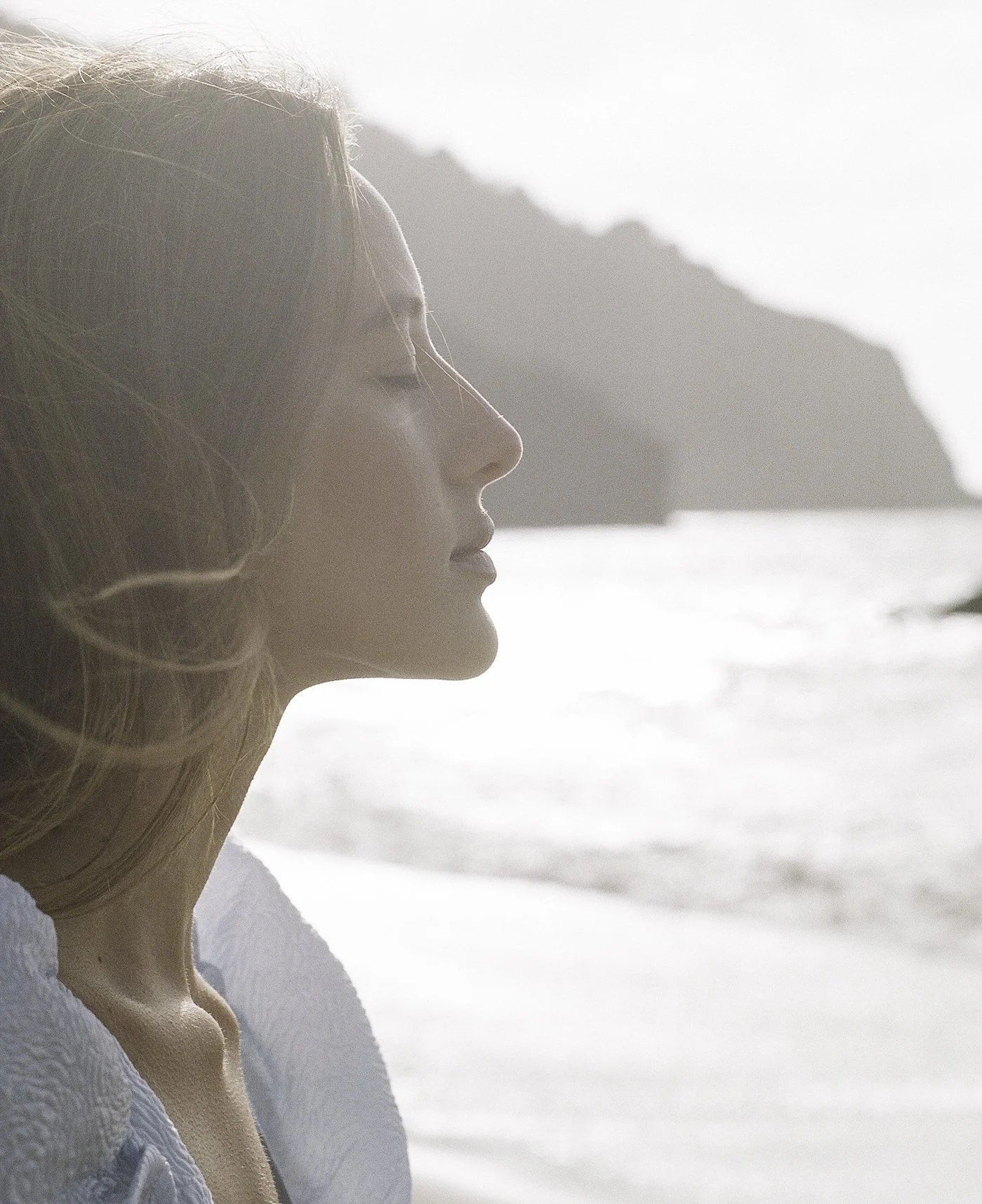 Editorial film portrait — woman in Cecilie Bahnsen blue organza dress in profile, hair blown by wind, golden coastal backlight dissolving background, by Radostina Boseva