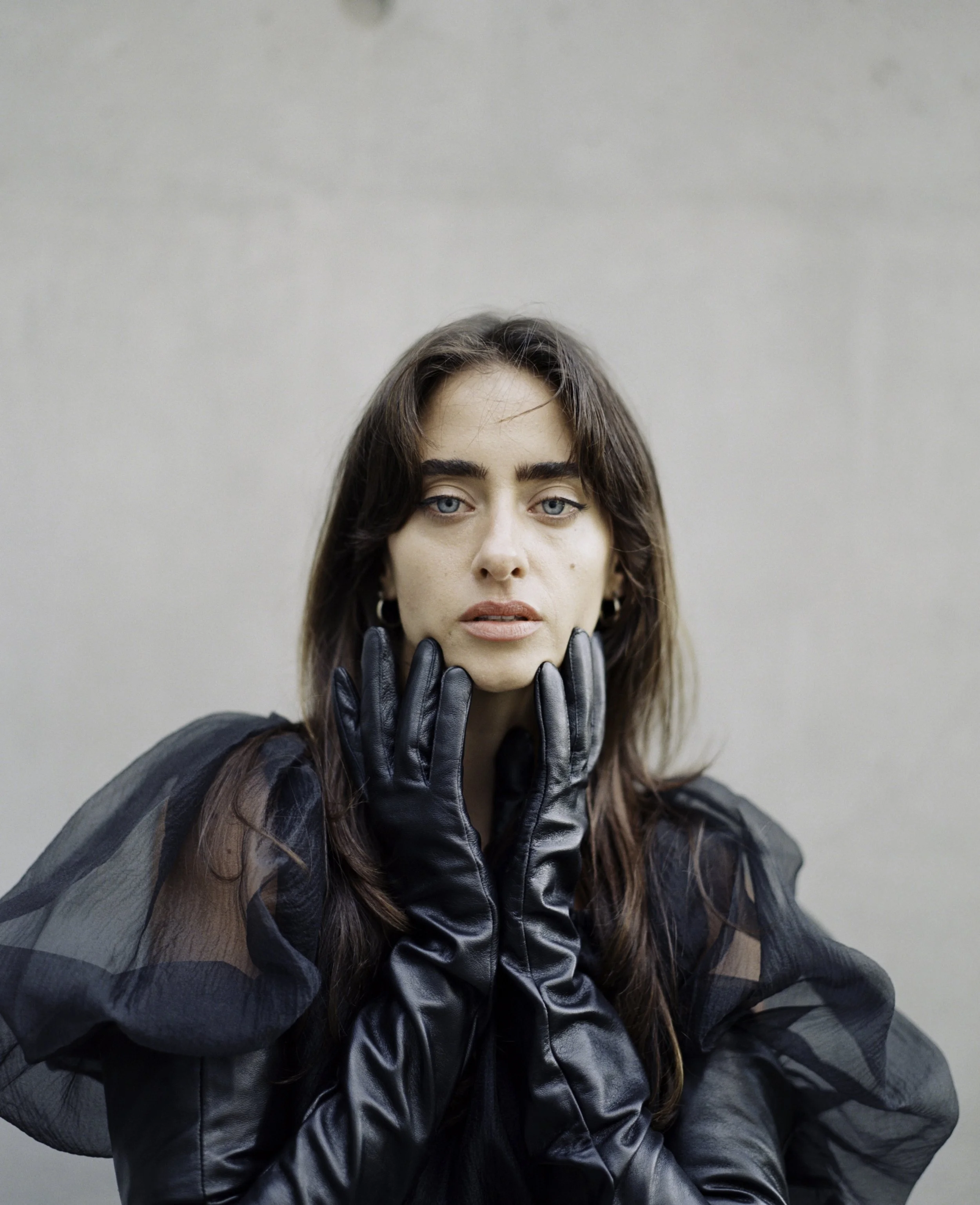 Fashion editorial portrait on film — woman in black organza dress and long leather gloves, hands framing face, shot against grey wall by Radostina Boseva