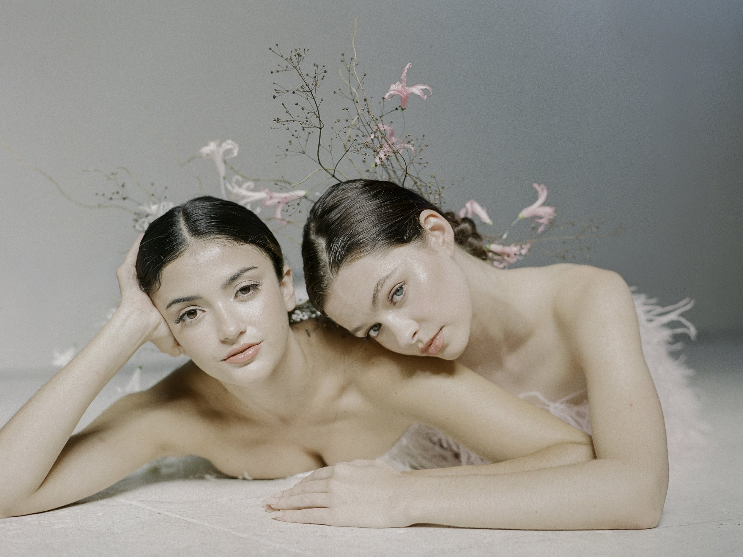 Fine art editorial film portrait — two women lying on studio floor with pink lilies and dried branches woven into their hair, both looking directly at camera, by Radostina Boseva
