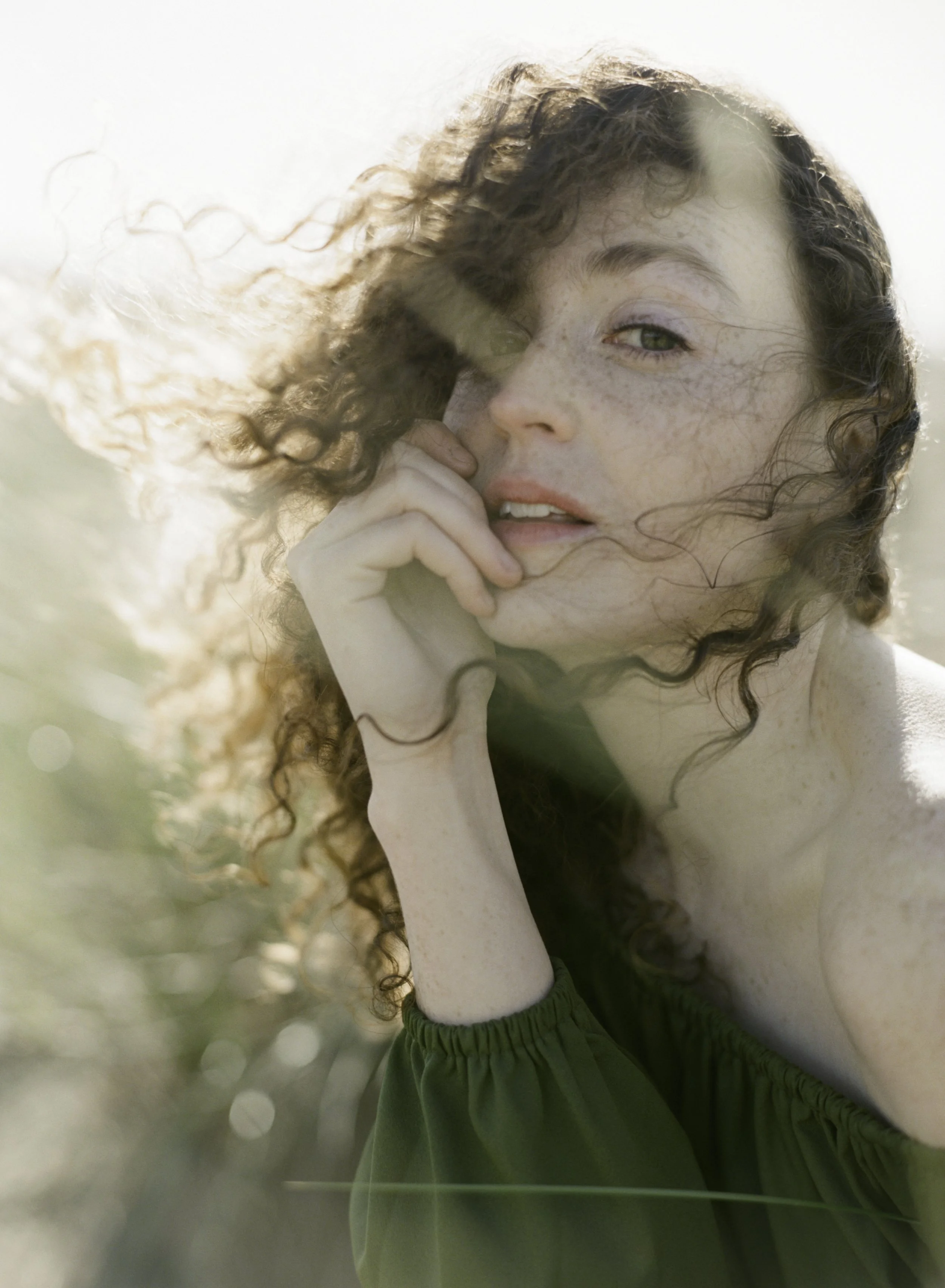 Intimate editorial film portrait of woman with curly auburn hair and freckles in green dress, hand to chin, warm lens flare, by Radostina Boseva