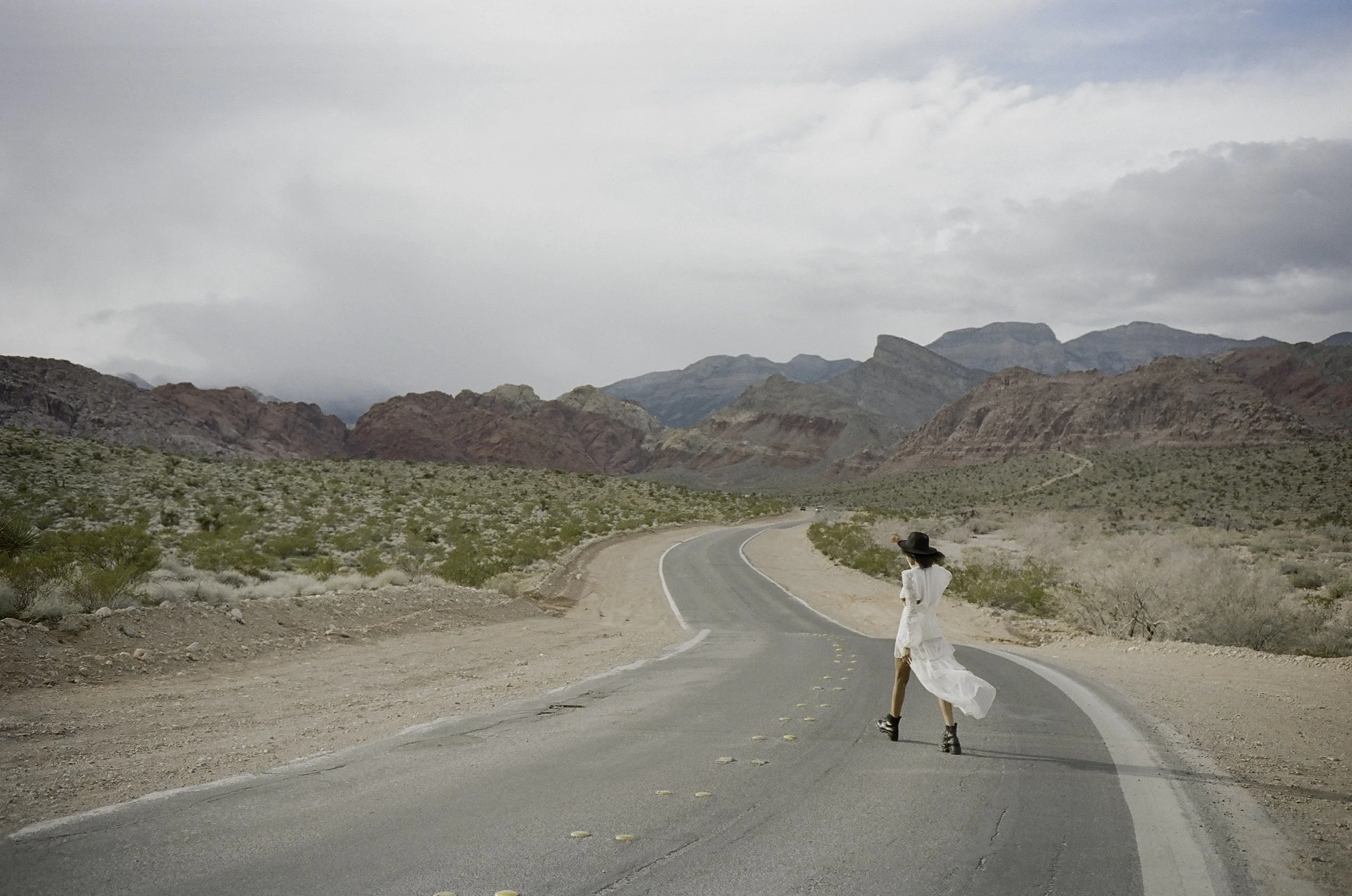 Editorial fashion film photograph — woman in white lace dress and black hat crossing empty desert highway, Red Rock Canyon mountains stretching behind her, Nevada, by Radostina Boseva