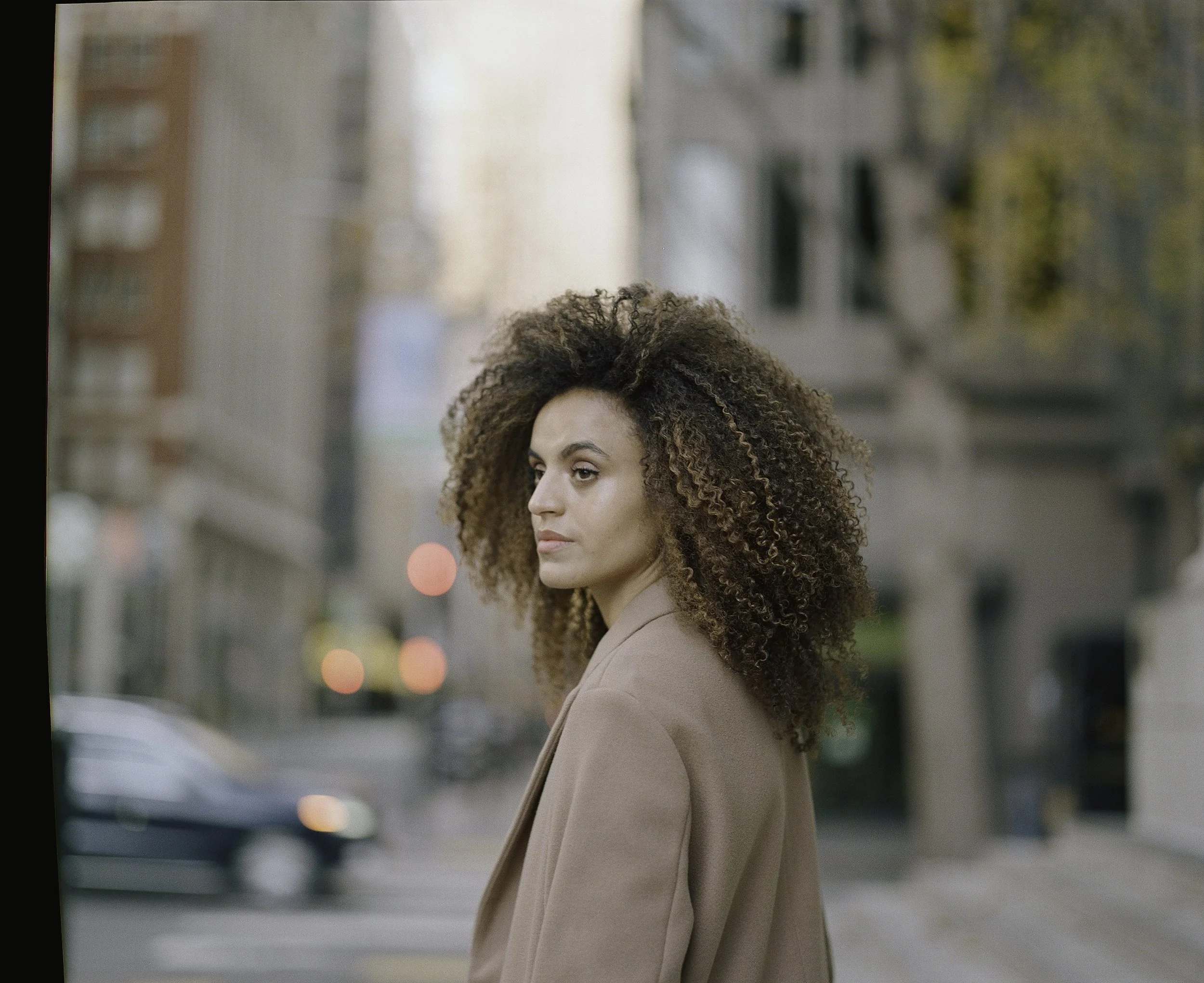 Editorial street portrait on film — woman with natural afro in camel coat turning to look over her shoulder on city street, red traffic lights in autumn bokeh behind her, by Radostina Boseva