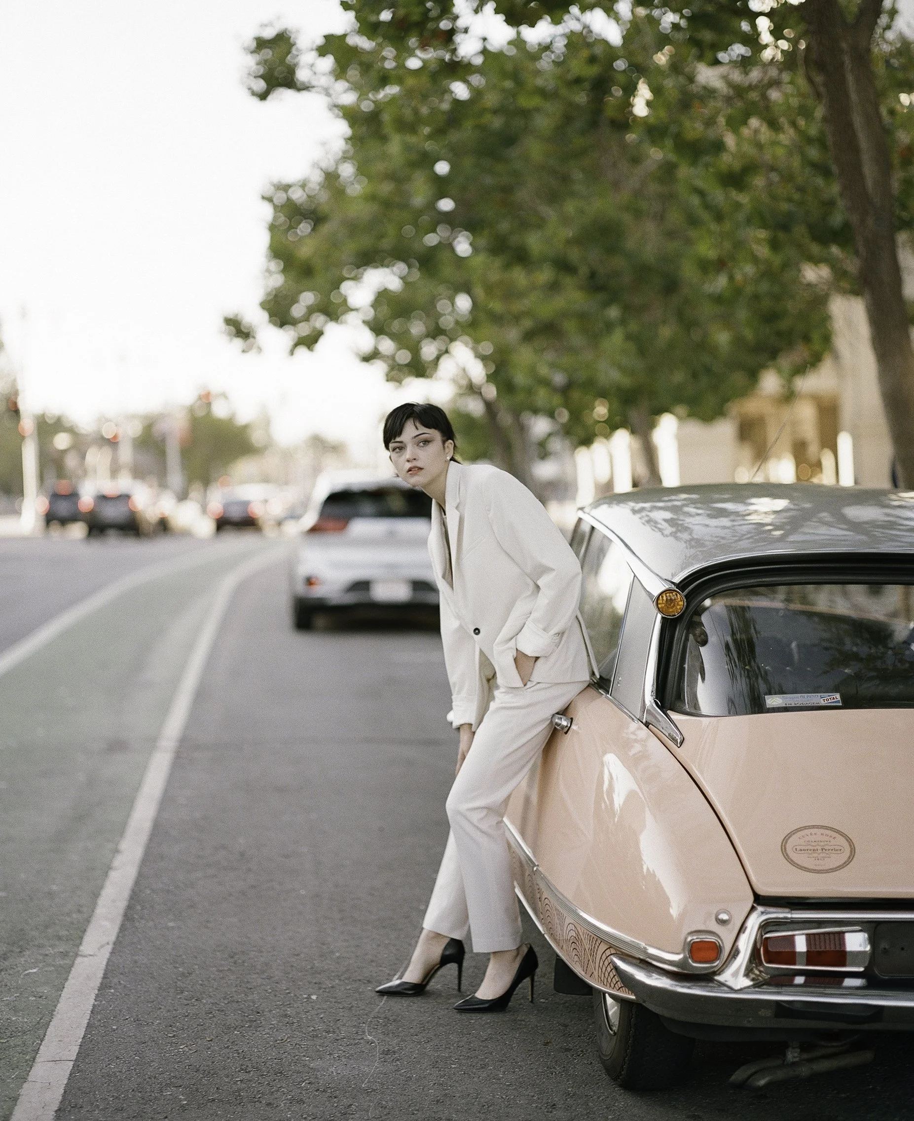 Editorial fashion portrait on film — dark-haired woman in cream suit leaning against pink Citroën DS on city street at dusk, direct gaze, by Radostina Boseva