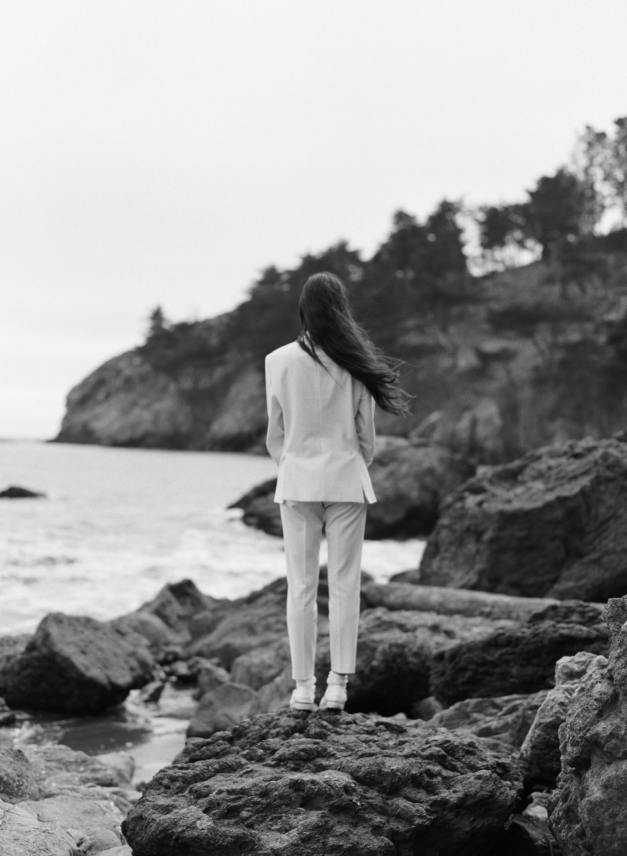 Editorial film photograph of woman in white suit standing with back turned on rocky California coastline, hair blown by wind, by Radostina Boseva