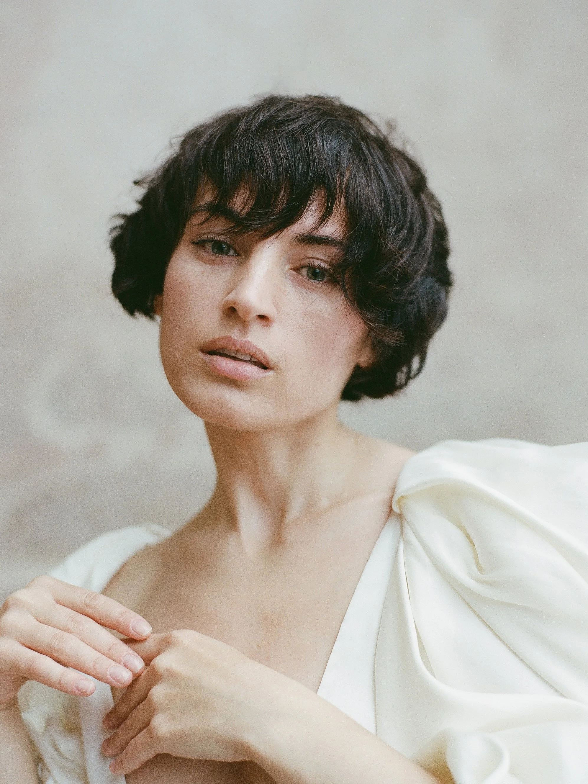 Intimate editorial film portrait — dark-haired woman in cream draped dress, hands at chest, direct gaze, shot on analogue film by Radostina Boseva